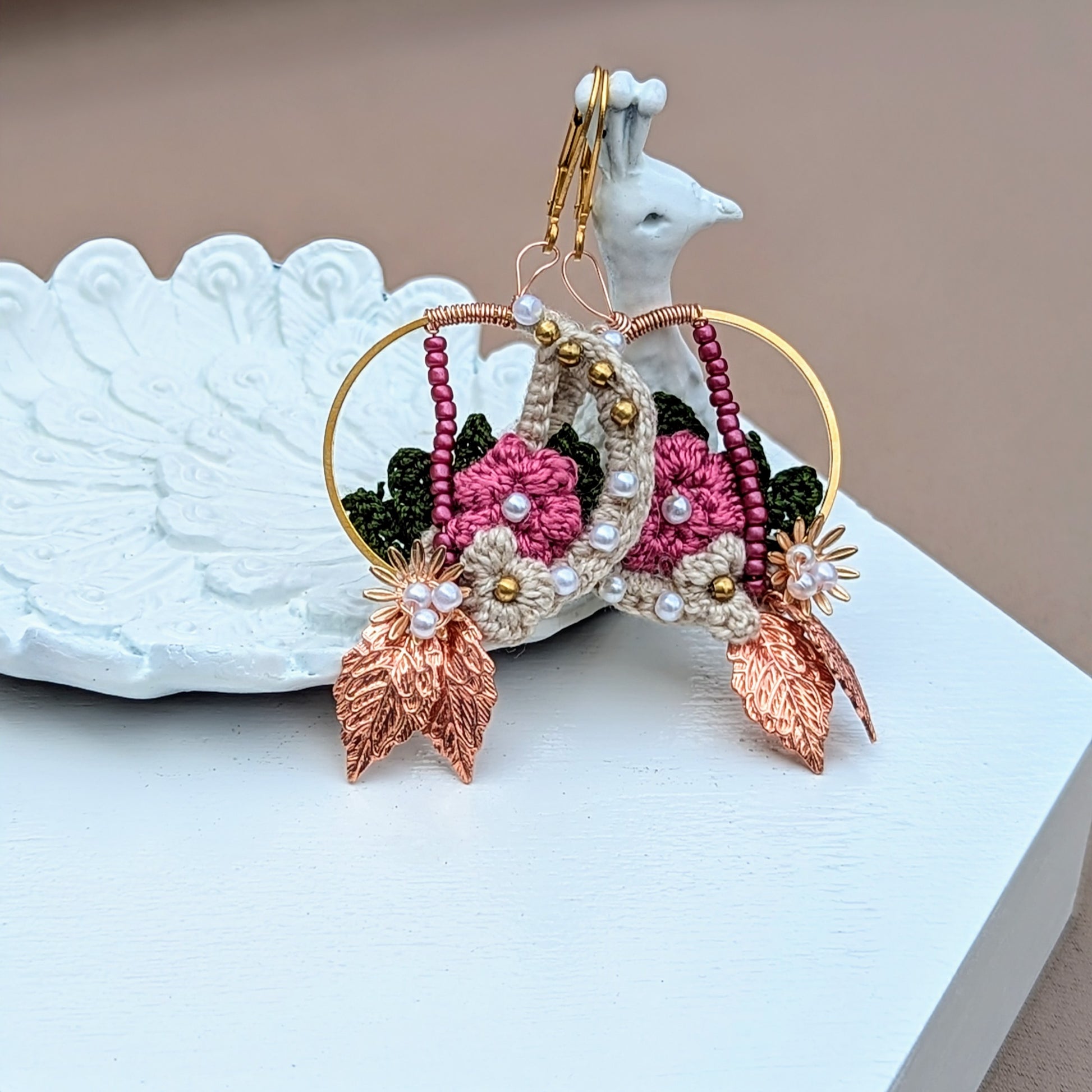 Pictured on a white peacock trinket dish, this handcrafted Indian-inspired earrings by Mayaani Jewellery, a UK sustainable brand features cream, pink, and dark green floral and leaf details. Adorned with solid brass, pink, and seed pearl beads, these earrings are finished with rose gold leaves and gold-plated stainless steel clasps. Lightweight and stunning, they're ideal for any occasion from weddings to everyday wear.