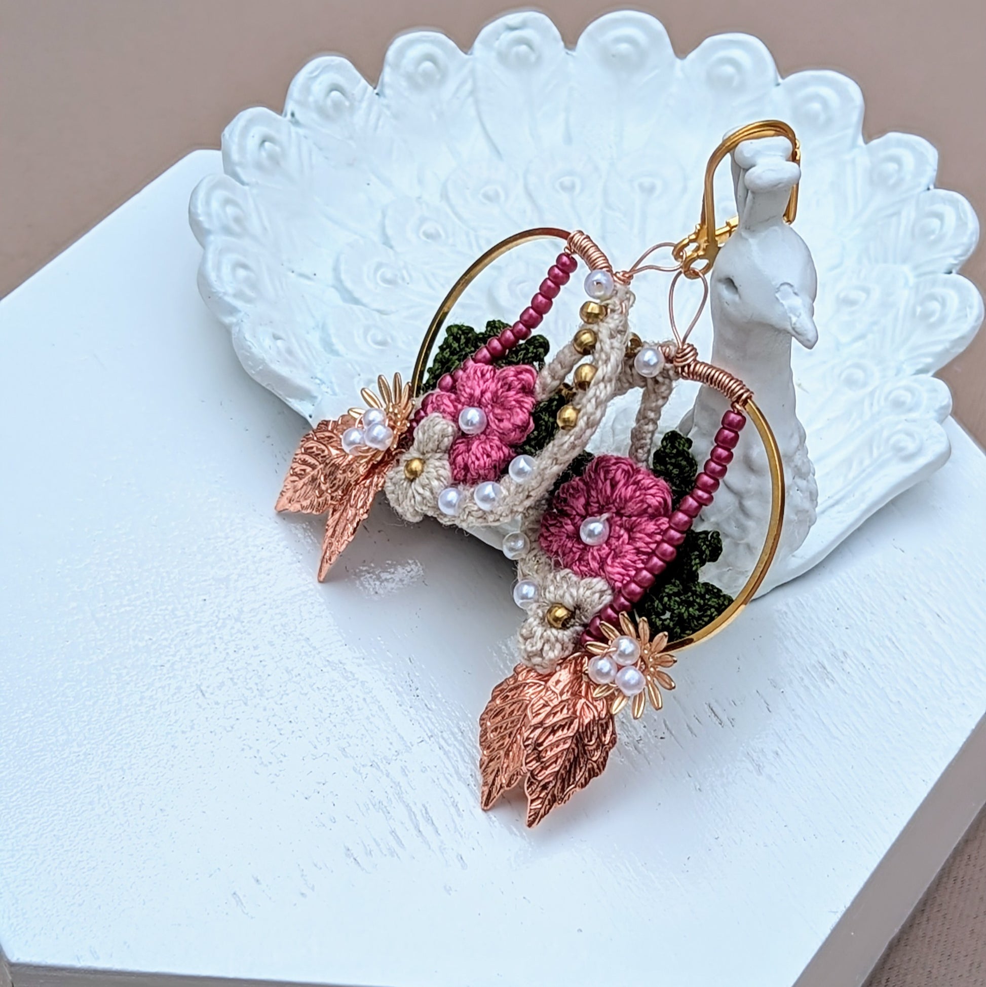 Delight in handcrafted Indian-inspired earrings by Mayaani Jewellery, a UK brand focused on sustainability. Pictured on a peacock trinket dish they feature cream, pink, and dark green floral and leaf motifs, enhanced by solid brass, pink, and seed pearl beads. Finished with rose gold leaves and gold-plated stainless steel clasps, they are lightweight and perfect for everything from weddings to everyday wear.