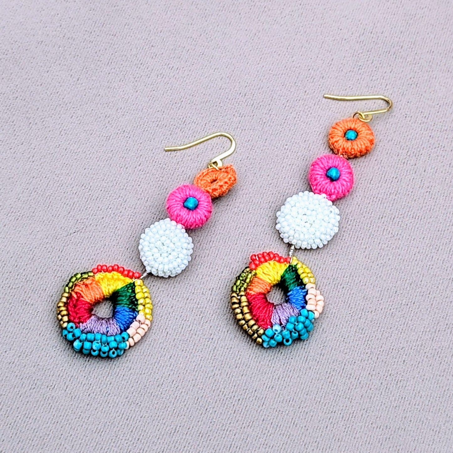 Discover a stunning pair of Indian-inspired rainbow earrings by Mayaani Jewellery UK. Featuring a cheerful rainbow crochet circle adorned with red, green, yellow, peach, gold, and blue hand beadwork, these earrings are framed by a white crochet and beaded design. The addition of pink and orange crochet accents enhances their charm, while gold-plated stainless steel hooks ensure lightweight comfort for all-day wear.