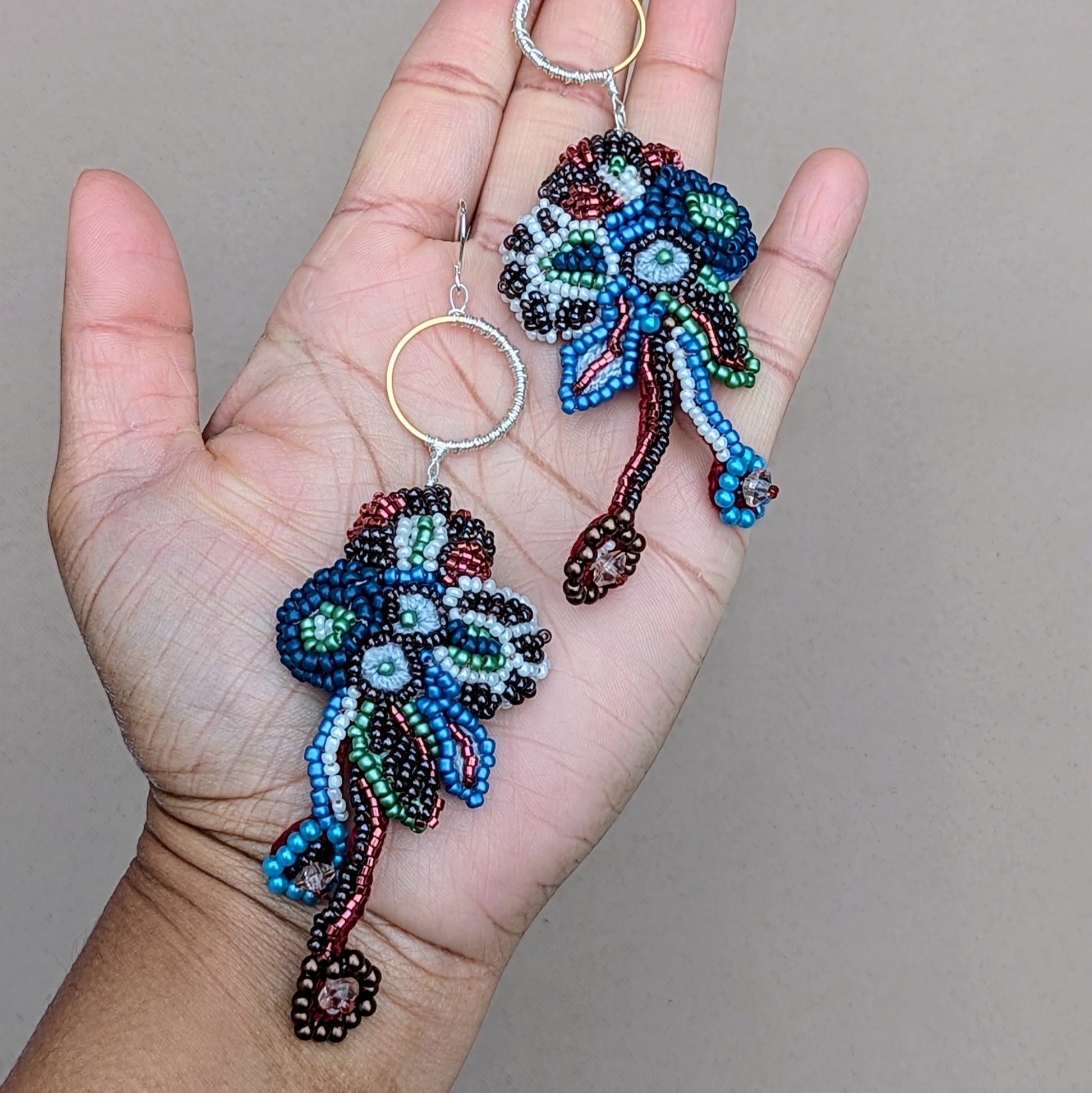 This image shows a unique, handcrafted pair of Indian-inspired earrings. Adorned with colourful beadwork in floral shapes, they hang from silver hooks with gold accents. Exclusive to Mayaani Jewellery UK, they showcase exquisite craftsmanship and cultural artistry.