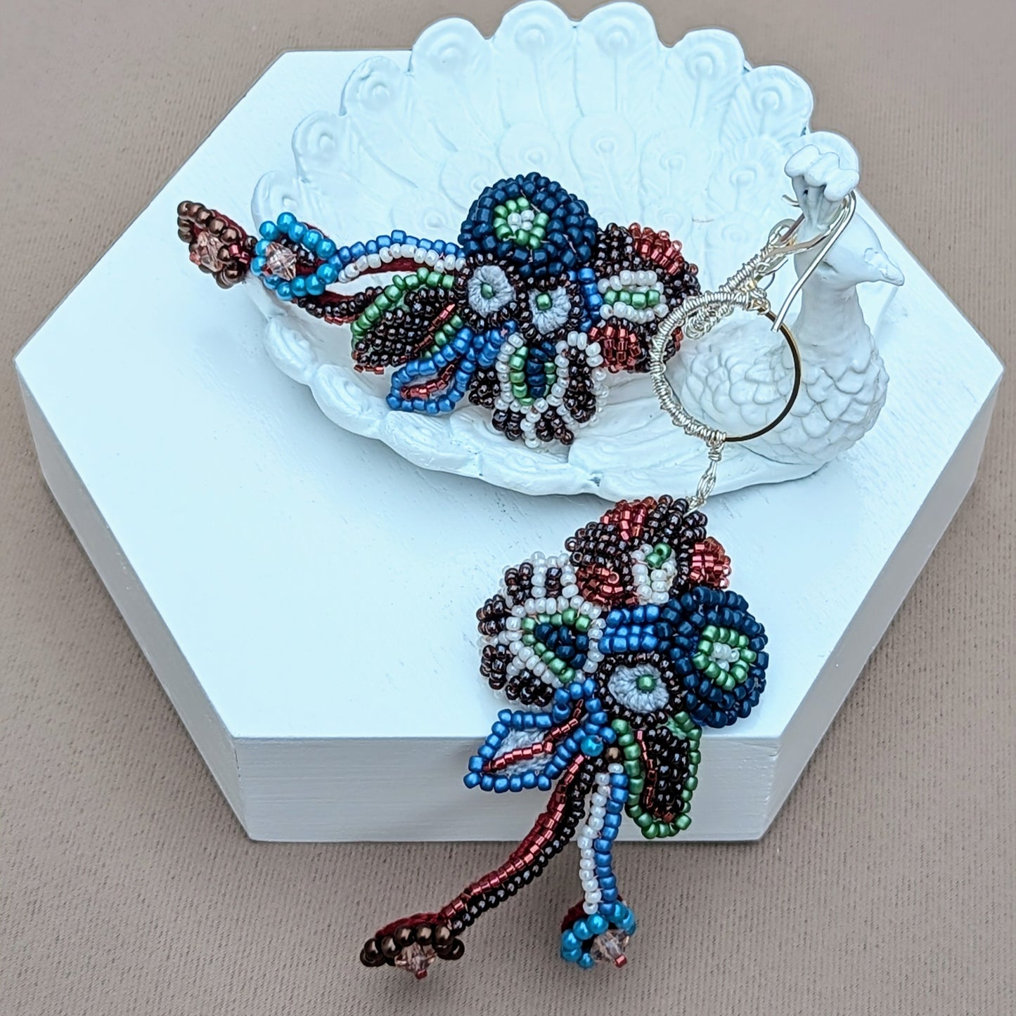 A pair of Indian-inspired earrings with intricate beadwork in floral patterns hanging from silver hooks displayed on a white peacock trinket dish. Exclusively from Mayaani Jewellery UK, this unique design showcases colourful craftsmanship. blending cultural inspiration with artistic beauty.