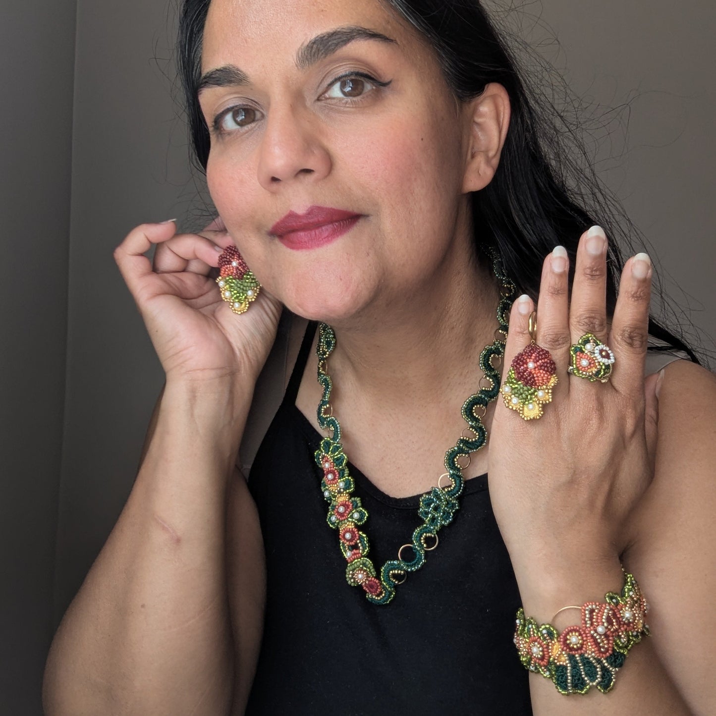 Mayaani Jewellery artisan Megha holding a pair of Rukmini earrings whilst modelling the Rukmini 5-in-1 Indian hand bracelet worn as separate elements including a cocktail ring, statement bracelet and intricate necklace in red, emerald green, gold, yellow and olive green.