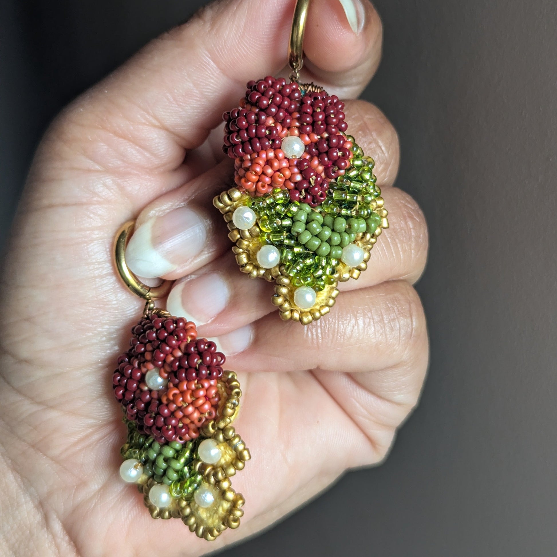 Hand holding a pair of dangle drop style Rukmini floral inspired earrings designed to complement the Rukmini statement Indian couture bridal occasion set by Megha of Mayaani featuring brick reds, peach tones, yellows, greens, gold and pearl accents.