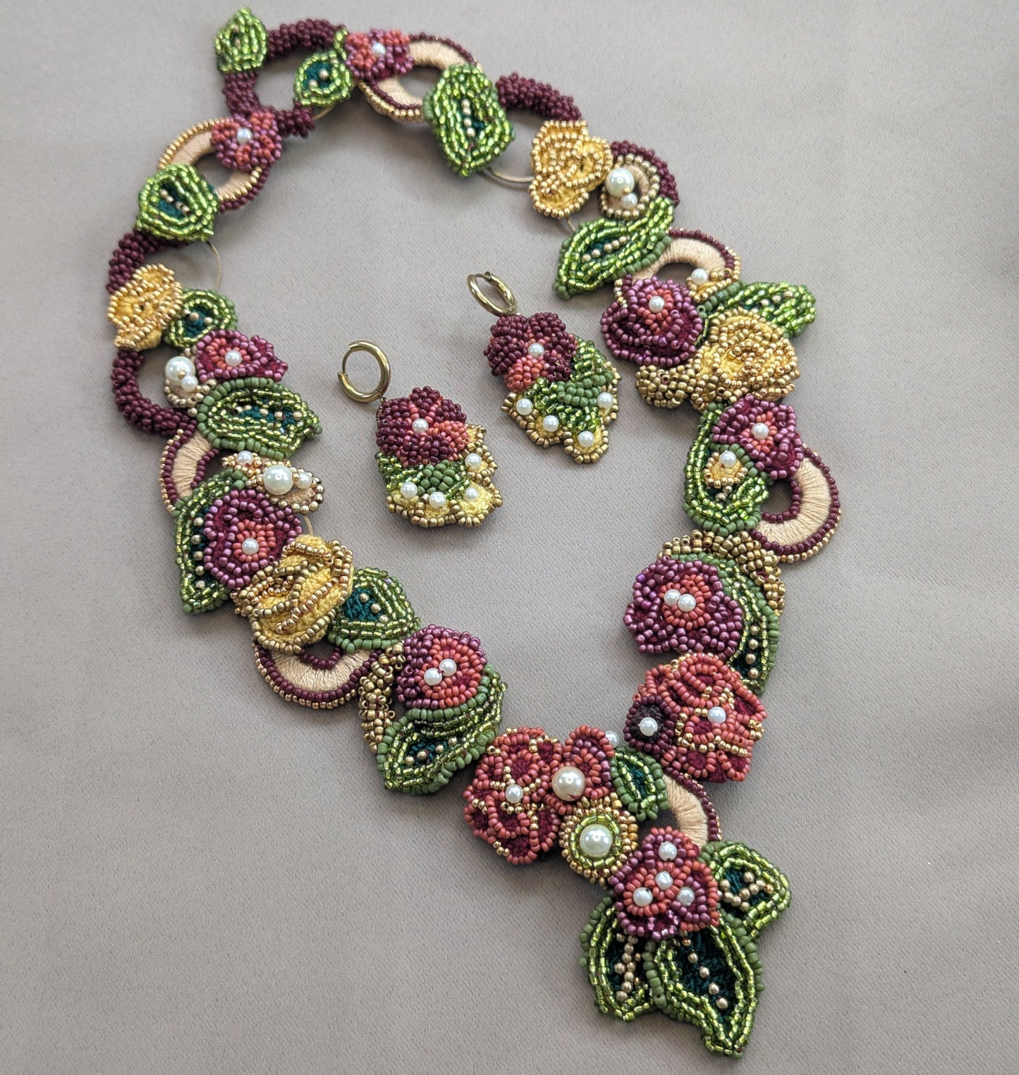 Flat lay view of the Rukmini 2-in-1 bead woven sustainably handcrafted designer necklace in red, green, yellow and gold with pearl accents surrounding matching Rukmini earrings on a neutral grey backdrop.