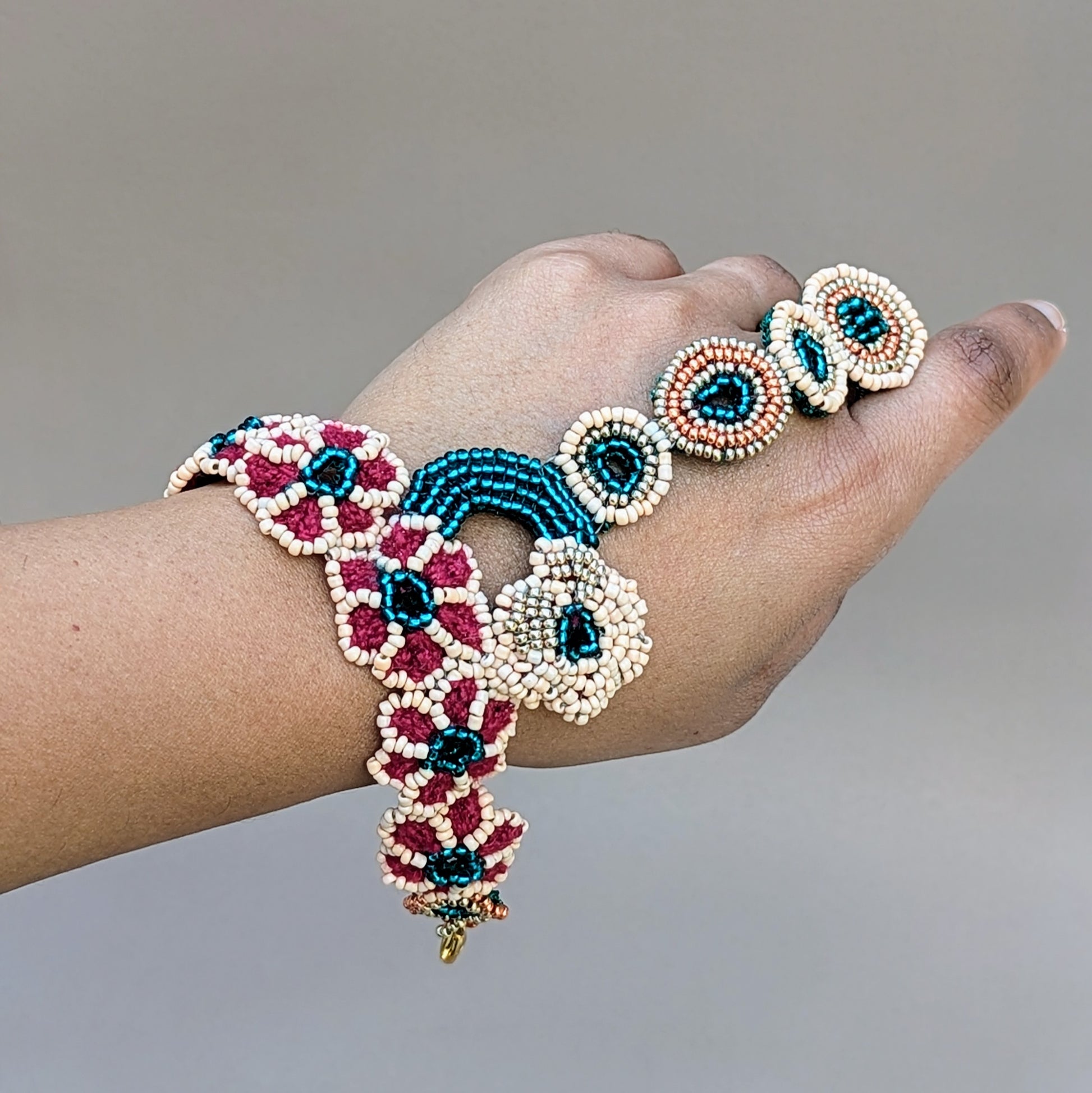 Beautiful haath phool, modelled on a hand, by sustainable UK brand Mayaani Jewellery, combining traditional Indian aesthetics with sustainable luxury. This hand bracelet features emerald green, peach, rose gold and gold beads in circular and floral motifs, meticulously crafted with hand beadwork, crochet and wire wrapping techniques. Connecting the wrist to a finger, it's ideal for brides or for making a statement at formal events. 