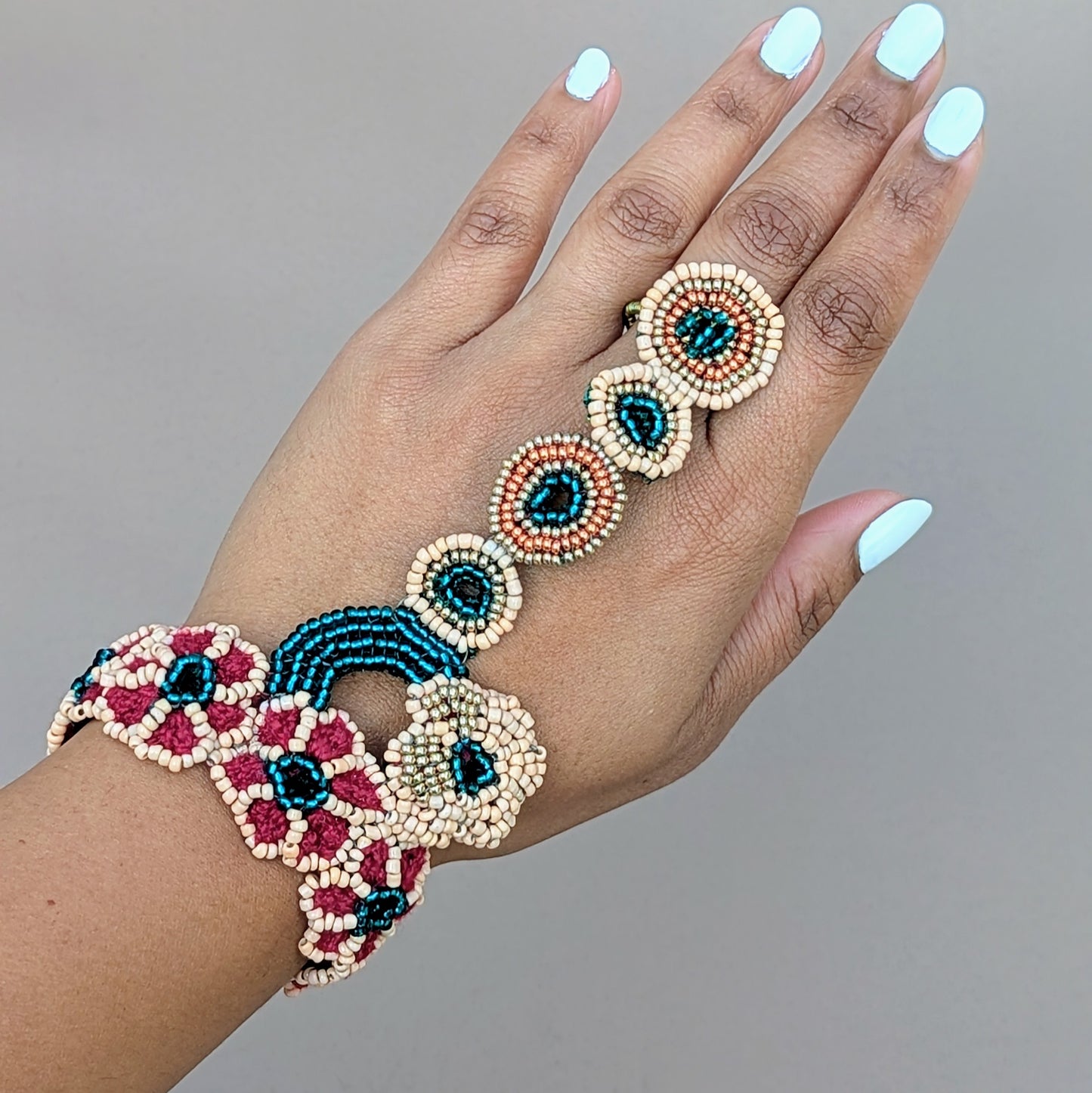 Hand showcasing a luxurious Indian hand bracelet or haath phool from sustainable brand Mayaani Jewellery UK, featuring a delicate chain of beaded rings in emerald green, peach, rose gold and gold tones. Intricately handcrafted with hand beadwork, crochet and wire work, the bracelet's floral and circular motifs create a stunning connection between wrist and finger. Designed to complement bridal attire or be worn independently for special occasions.