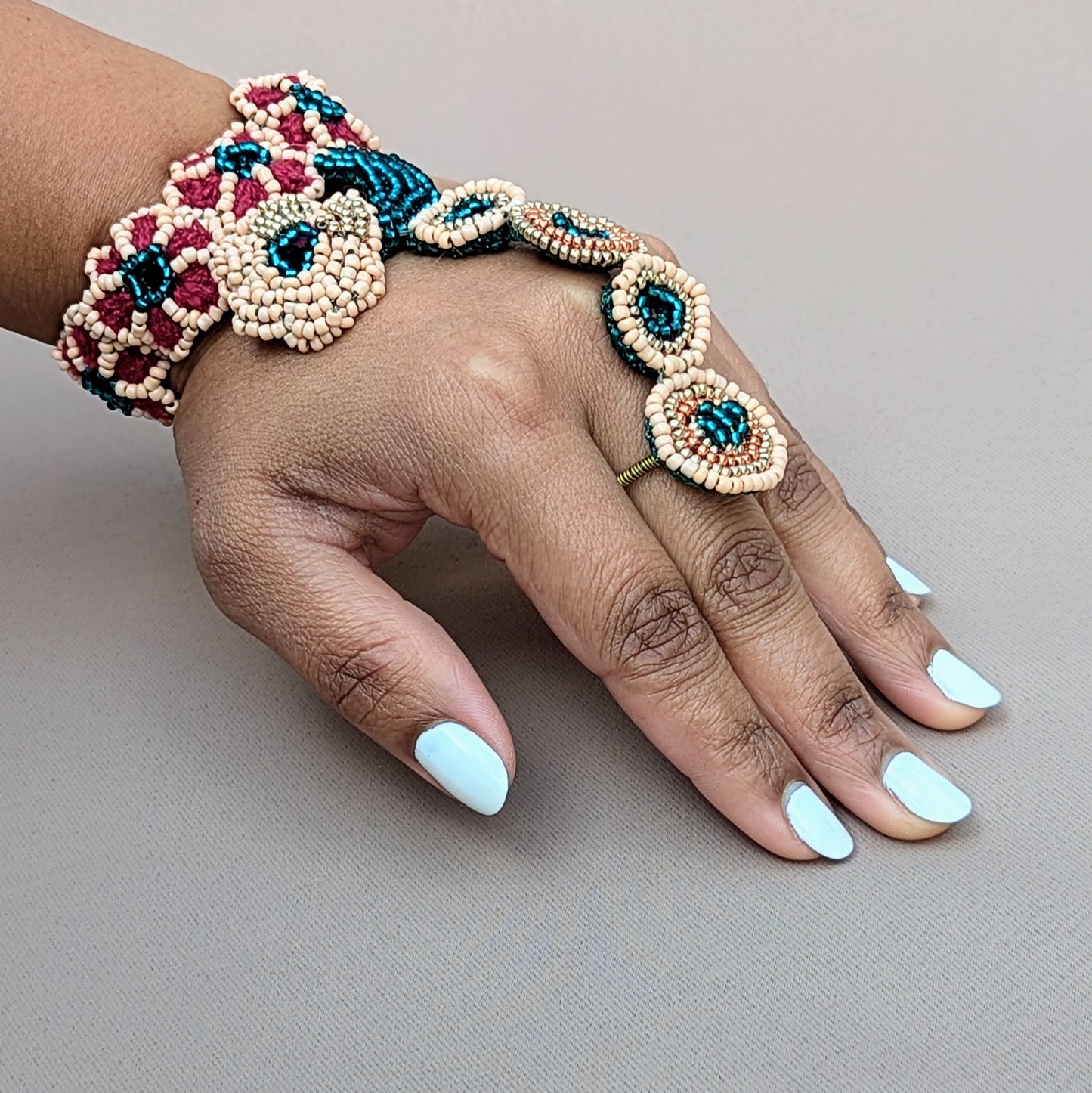 Hand modelling a handcrafted haath phool from sustainable brand Mayaani Jewellery UK, featuring intricate beadwork in emerald green, peach, rose gold and gold tones. The design connects the wrist to a finger with beaded rings and floral patterns. Created using hand crochet, hand beadwork and wire wrapping techniques, this luxurious hand bracelet complements both bridal attire and formal occasions, adding a touch of elegance.