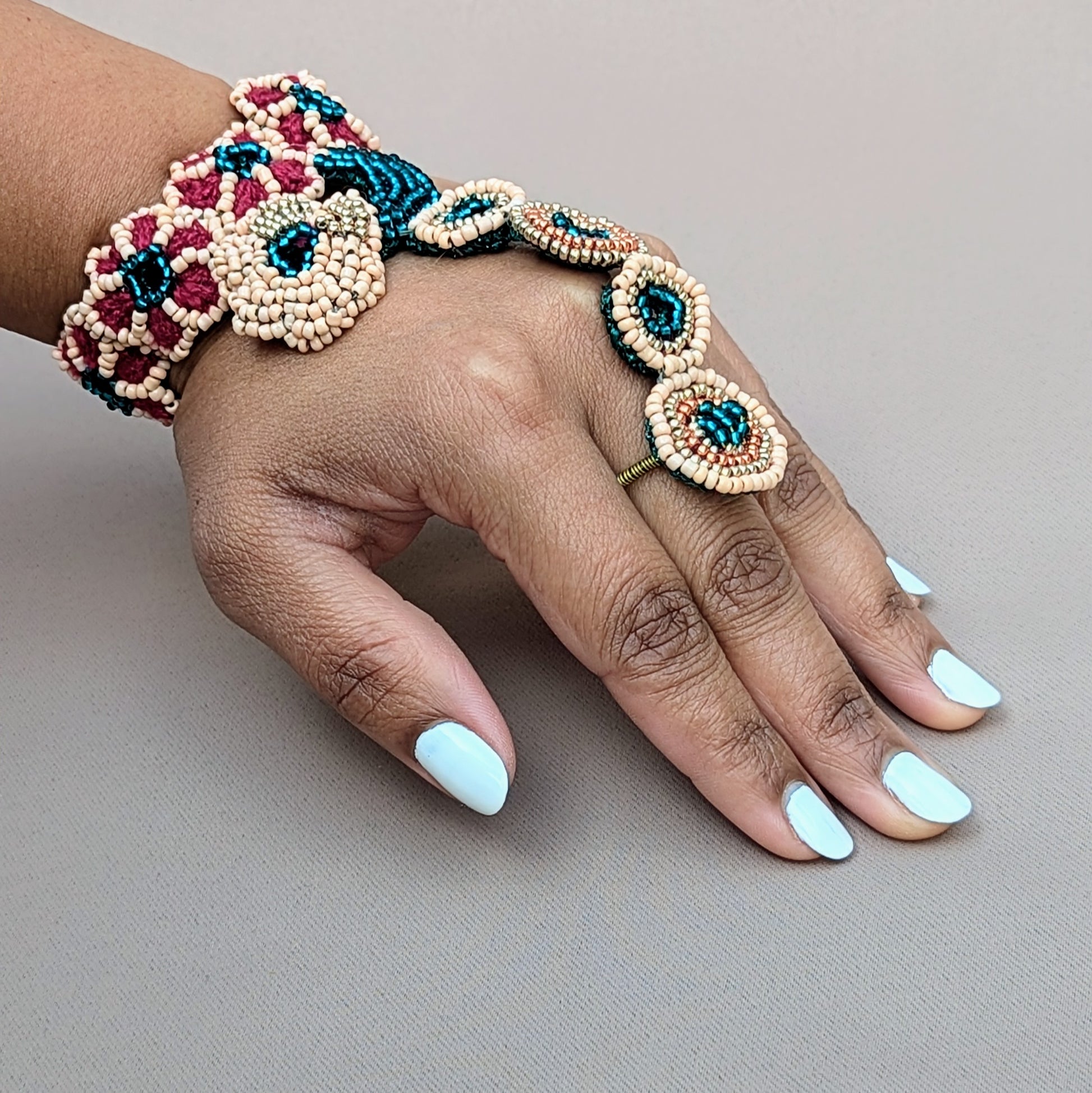 Hand modelling a handcrafted haath phool from sustainable brand Mayaani Jewellery UK, featuring intricate beadwork in emerald green, peach, rose gold and gold tones. The design connects the wrist to a finger with beaded rings and floral patterns. Created using hand crochet, hand beadwork and wire wrapping techniques, this luxurious hand bracelet complements both bridal attire and formal occasions, adding a touch of elegance.