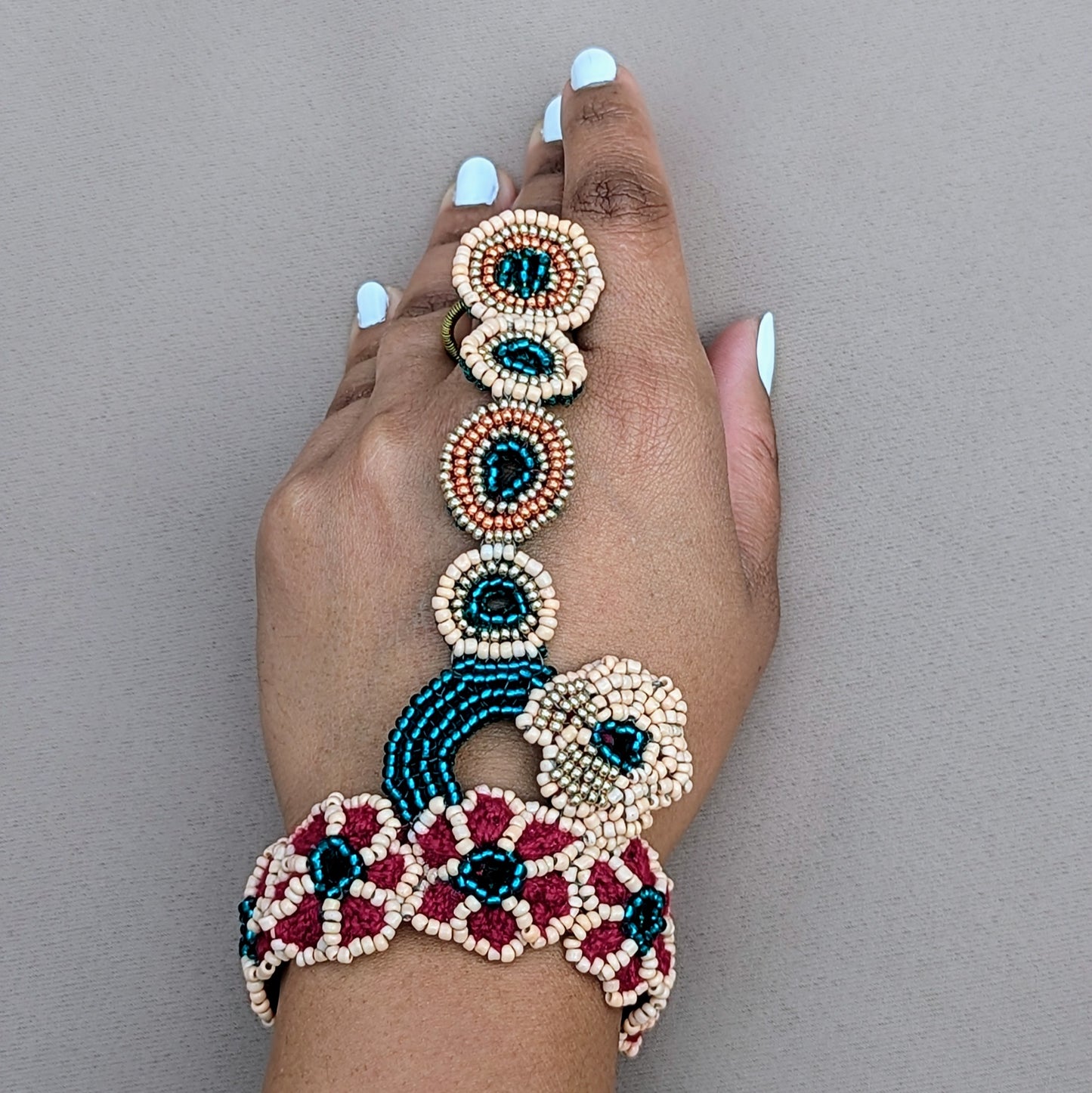 Elegant Indian haath phool handcrafted by Mayaani jewellery. This luxury hand bracelet features detailed beadwork in soft peach, emerald green, gold and rose gold tones, connecting the wrist to the finger with a series of floral and circular beaded motifs. A bold fashion piece, it combines traditional craftsmanship with modern flair, ideal for bridal wear or standalone use at special events.