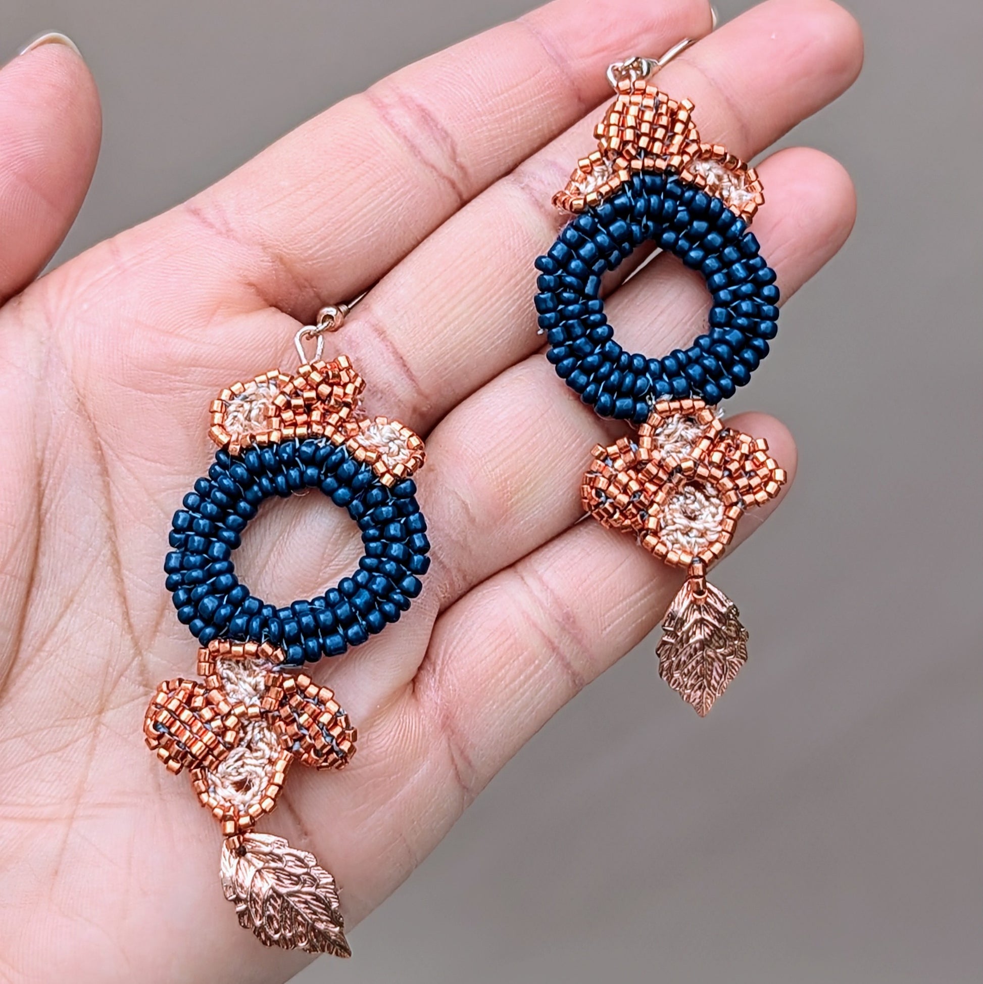 A beautiful pair of earrings with a large circular blue beaded centrepiece, complemented by floral motifs in copper Japanese Miyuki seed beads and golden leaves. The design incorporates delicate hand-crochet and beadwork, finished with rose gold-plated hooks. These handcrafted earrings from Mayaani Jewellery showcase a harmonious blend of deep blue glass seed beads and premium materials, reflecting sustainable fashion and artistry. Dimensions: max width 2.7cm, approx. length 7cm.