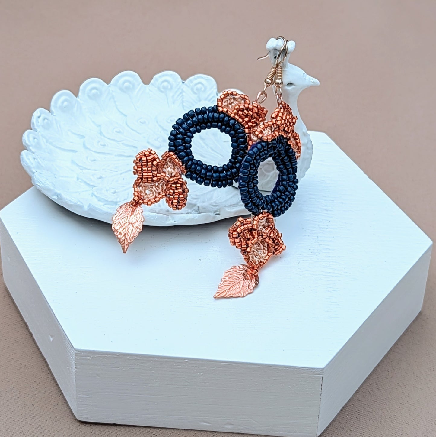 Showcasing a striking pair of earrings, these feature a large circular blue bead at the centre, surrounded by floral motifs created with copper Japanese Miyuki seed beads and accented with gold leaves. The exquisite design includes hand-crochet and beadwork, with rose gold-plated hooks providing a luxurious touch. Handcrafted by Mayaani Jewellery, these earrings highlight a blend of Indian inspiration and sustainable fashion. Dimensions: max width 2.7cm, approx. length 7cm.