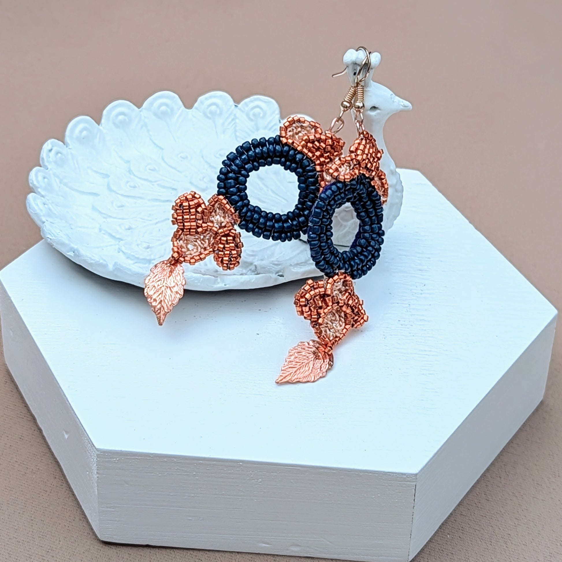 Showcasing a striking pair of earrings, these feature a large circular blue bead at the centre, surrounded by floral motifs created with copper Japanese Miyuki seed beads and accented with gold leaves. The exquisite design includes hand-crochet and beadwork, with rose gold-plated hooks providing a luxurious touch. Handcrafted by Mayaani Jewellery, these earrings highlight a blend of Indian inspiration and sustainable fashion. Dimensions: max width 2.7cm, approx. length 7cm.
