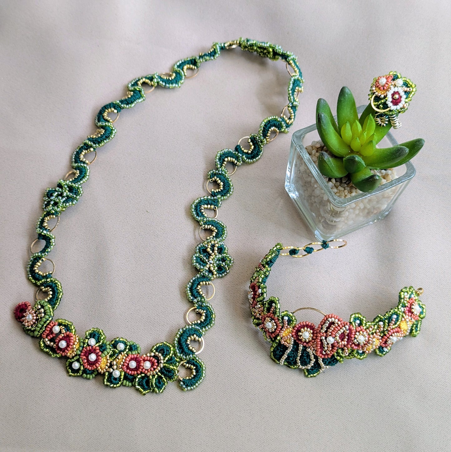 The Rukmini 5-in-1 Indian hand bracelet jewellery set featuring elements the bracelet can transform into including a necklace, statement bracelet, and adjustable cocktail ring with intricate green, red, pink, and yellow floral patterns, displayed on a light beige fabric background next to a small potted succulent.