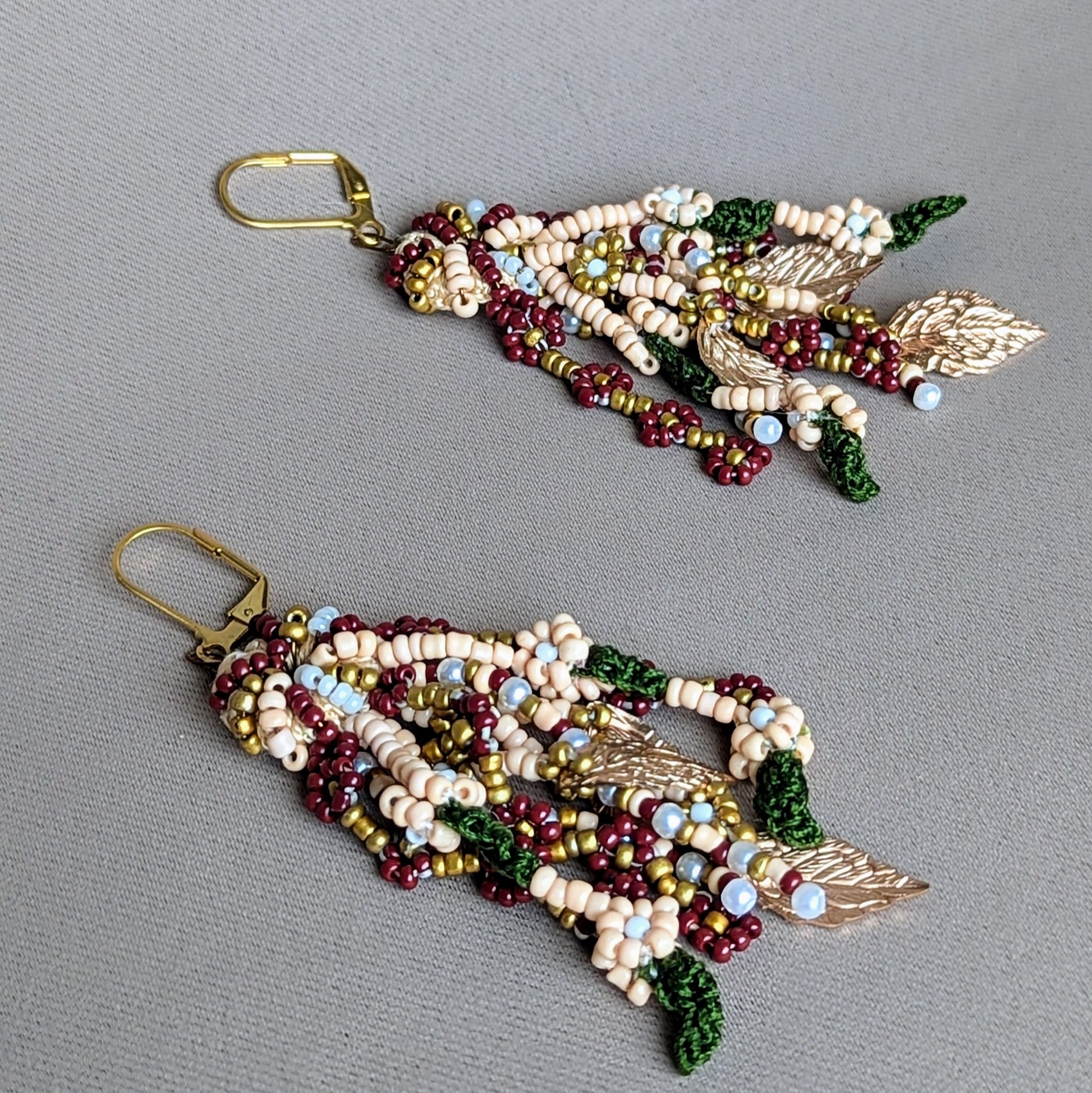 Close-up of handcrafted multi-strand Indian bridal occasion earrings by Mayaani Jewellery UK featuring red, green, gold and pearl seed beads with gold-plated leaves and lever back hooks. Photographed against a neutral grey background.