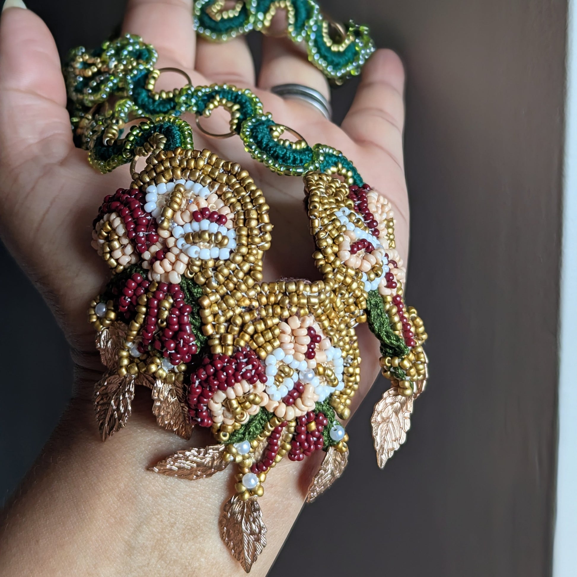 Hand holding the floral motif inspired Aishwarya green, gold, red, statement pendant with the Rukmini emerald green and gold chain exclusively handmade for Mayaani Jewellery UK.