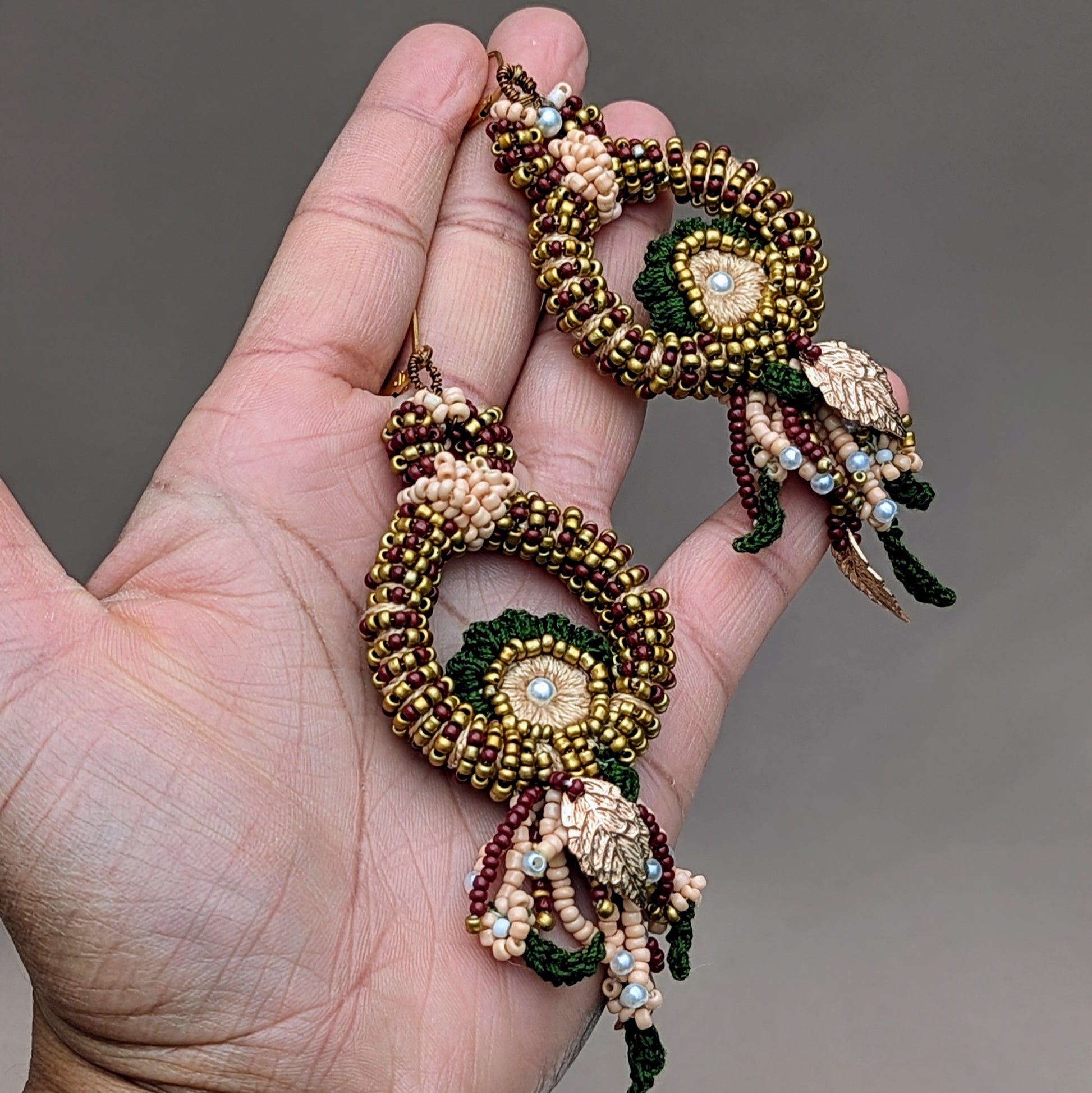 Hand held view of a pair of peacock shaped red, green, gold and pearl Aishwarya Indian artisan earrings handcrafted by Megha of Mayaani UK. 