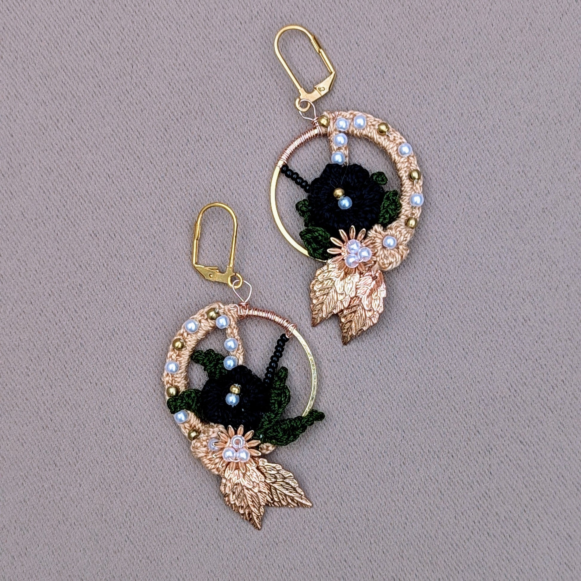 One-of-a-kind Aloki black and gold hoop earrings with floral motif, handcrafted in crochet using black and olive green threads, accented with gold-plated leaves, solid brass beads, and seed pearls, designed by Mayaani Jewellery UK.