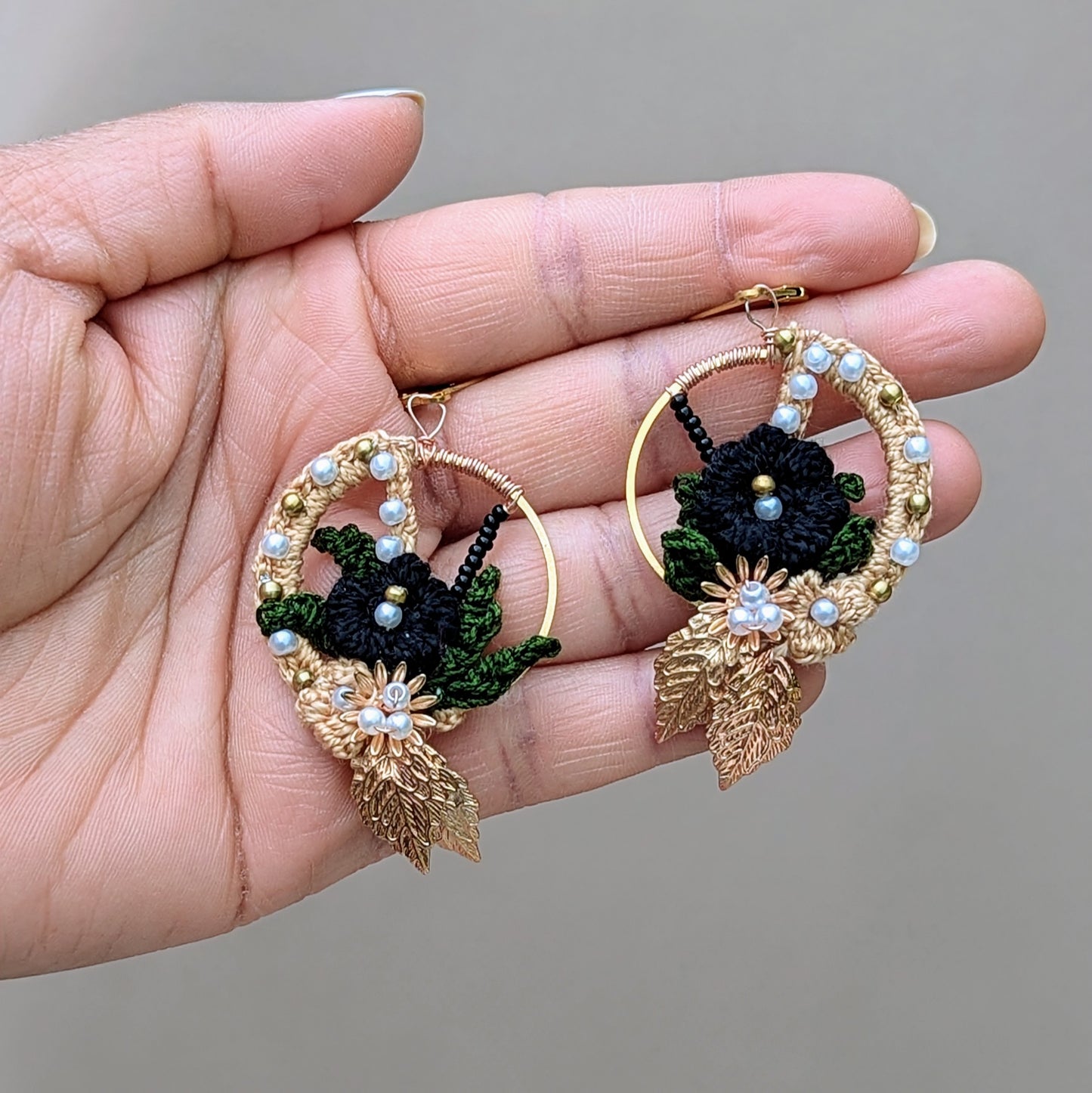 Hand holding Aloki black and gold hoop earrings, showcasing floral crochet design with seed pearls and golden leaves.