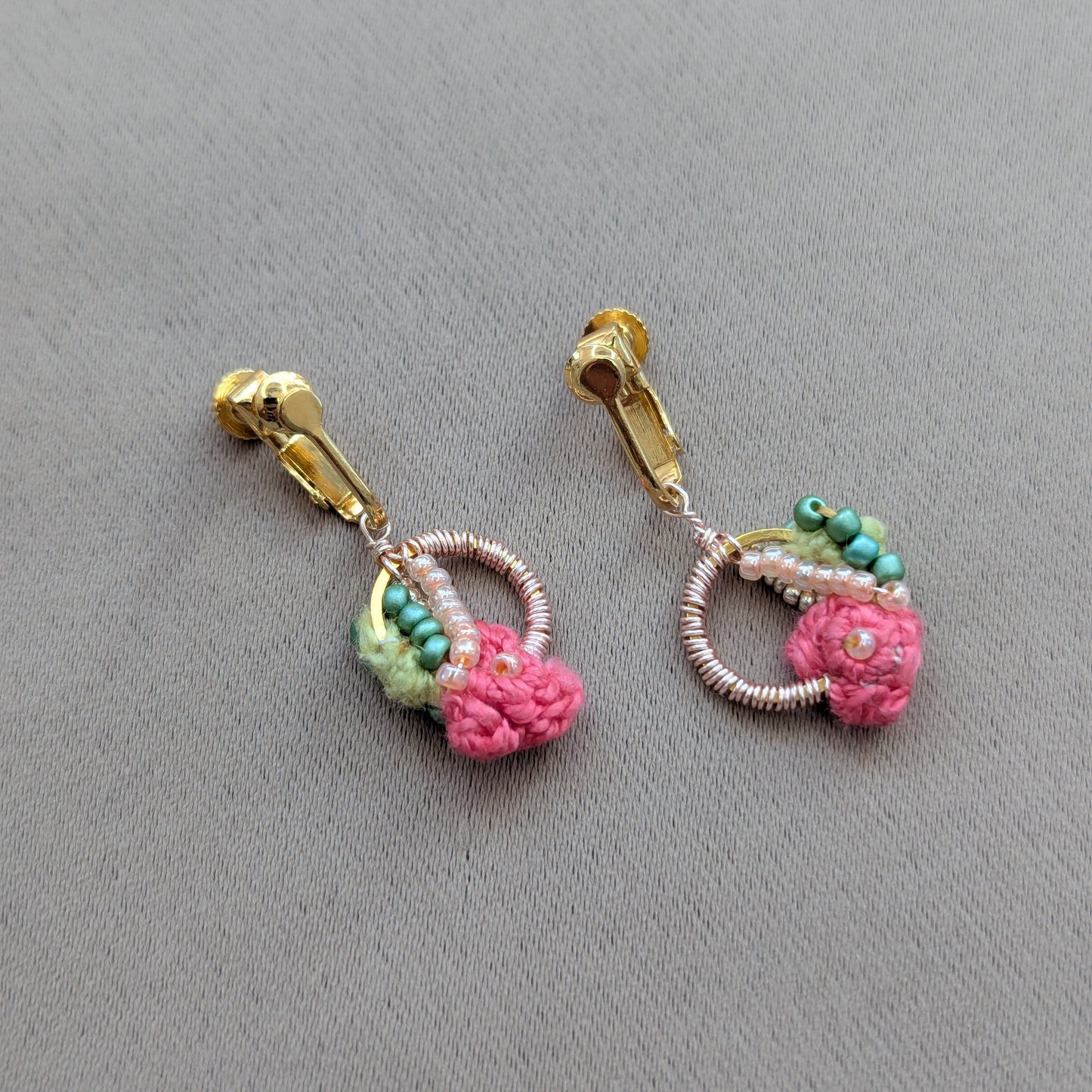 A pair of Anandi handmade gold and pink clip-on earrings featuring pink crochet flowers green bead accents, and a rose gold circular wire detail, displayed on a textured grey background.