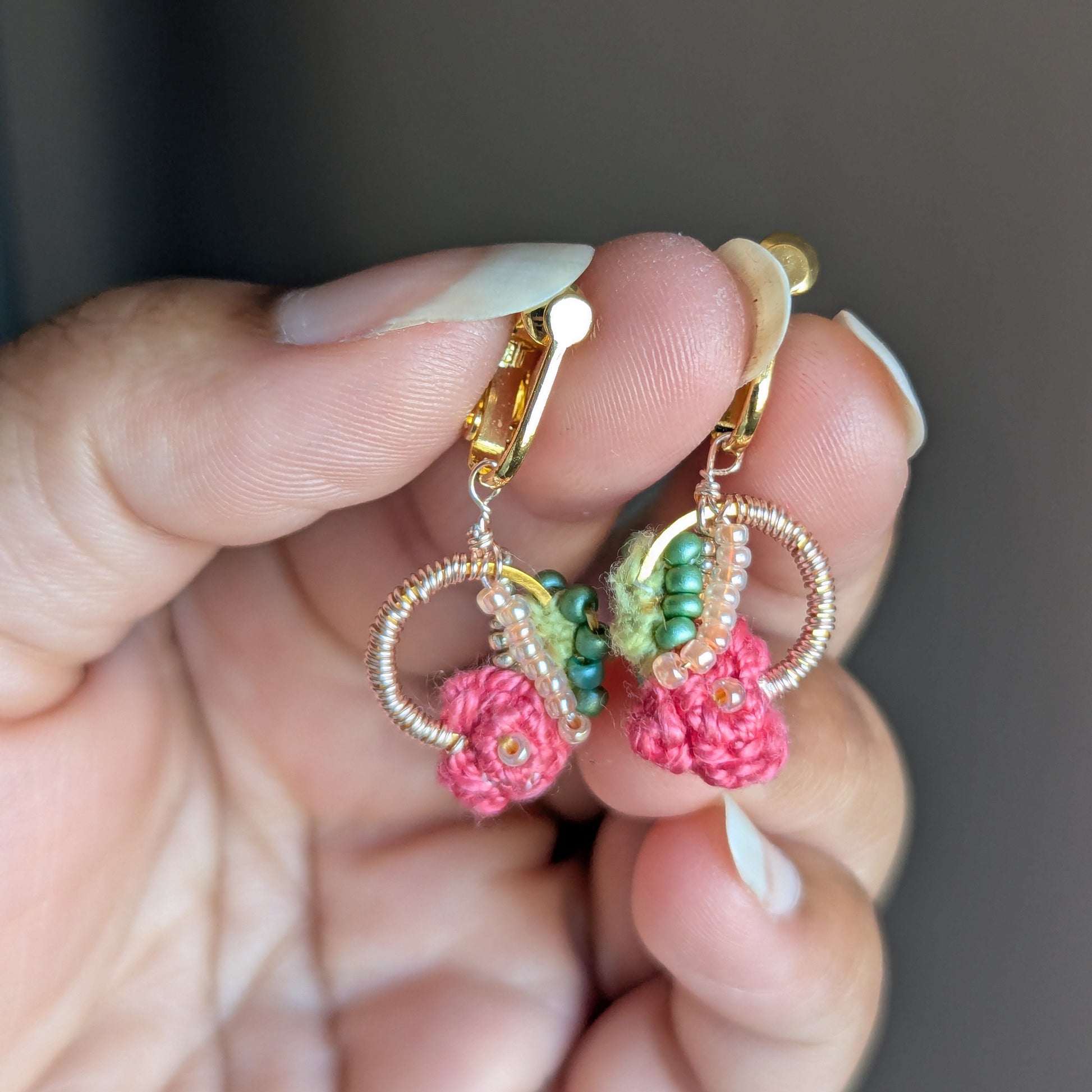 Close-up view of Anandi artisan made pink, green and gold dangle drop style earrings held gently between fingers against a neutral grey backdrop.