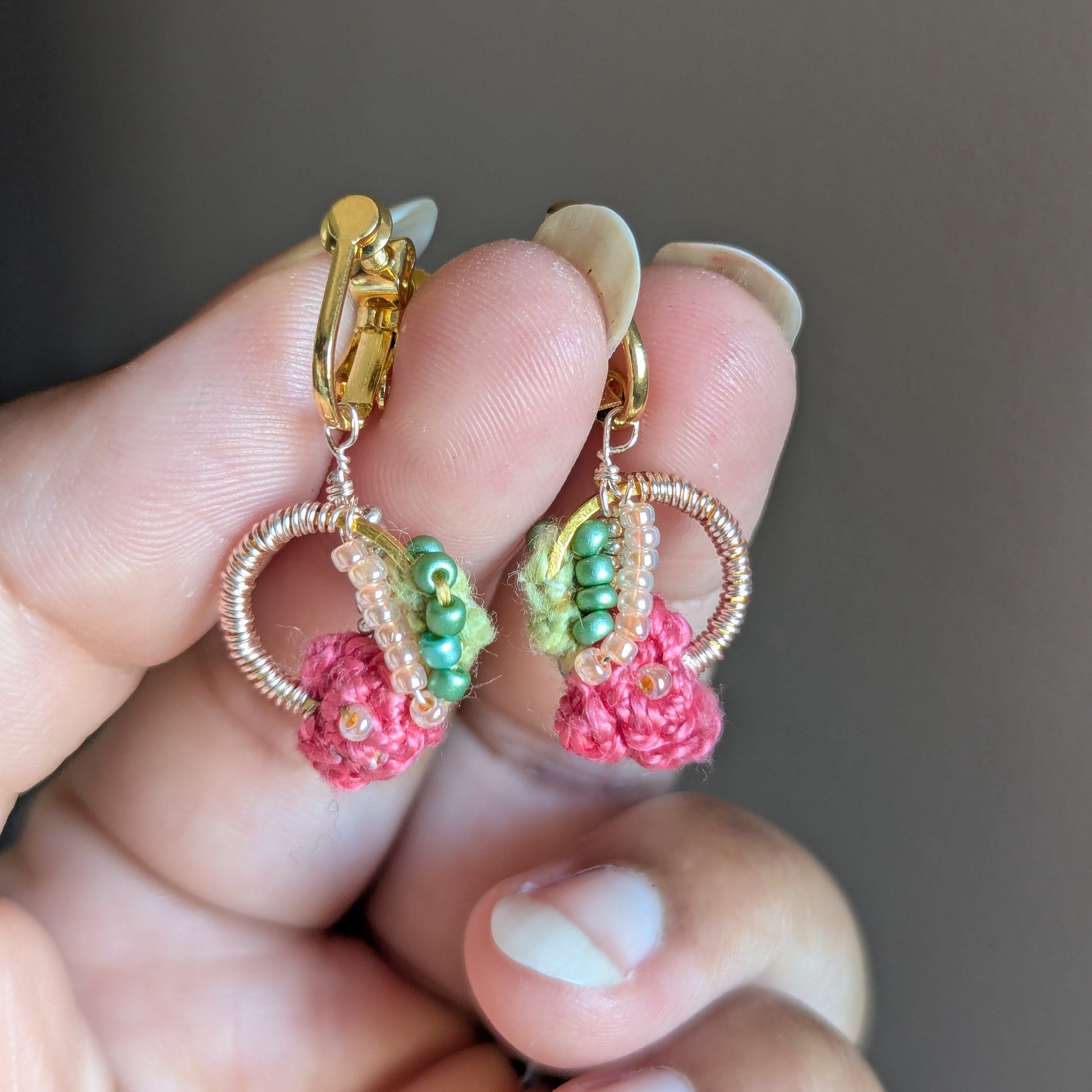 Close-up of hands holding the Anandi one-of-a-kind comfortable clip-on earrings by Mayaani UK featuring gold stainless steel screwback clasp, rose gold plated wire and pink and green crochet and bead details.