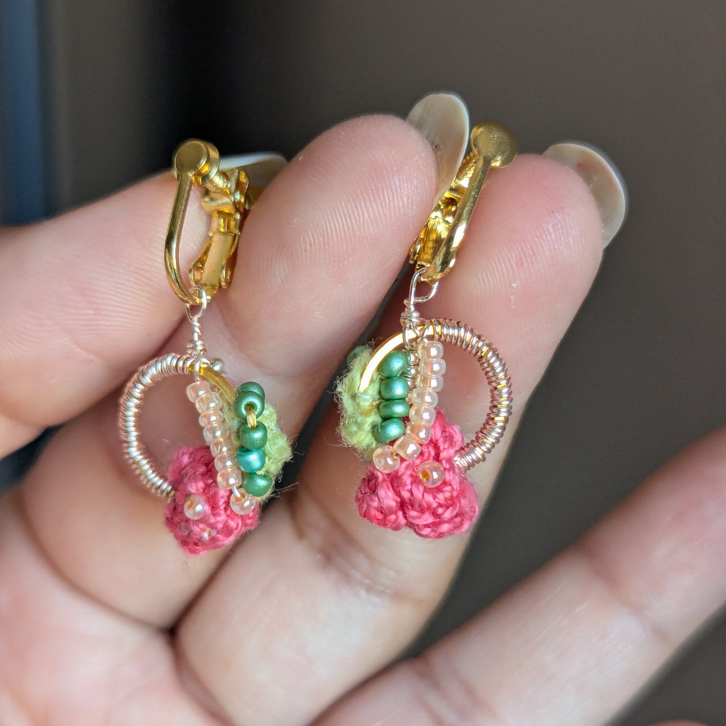 The Anandi pair of handmade drop style earrings featuring gold hoops with pink crochet flowers, green seed bead details, and small pearl-like accents, held in a person's hand.