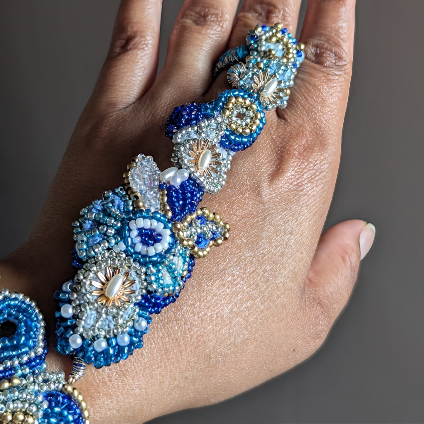 Hand wearing the Nira 5-in-1 beaded floral ring and matching beaded bracelet. The jewellery features blue, white, and gold seed beads in a detailed floral pattern, set against a solid dark grey background.