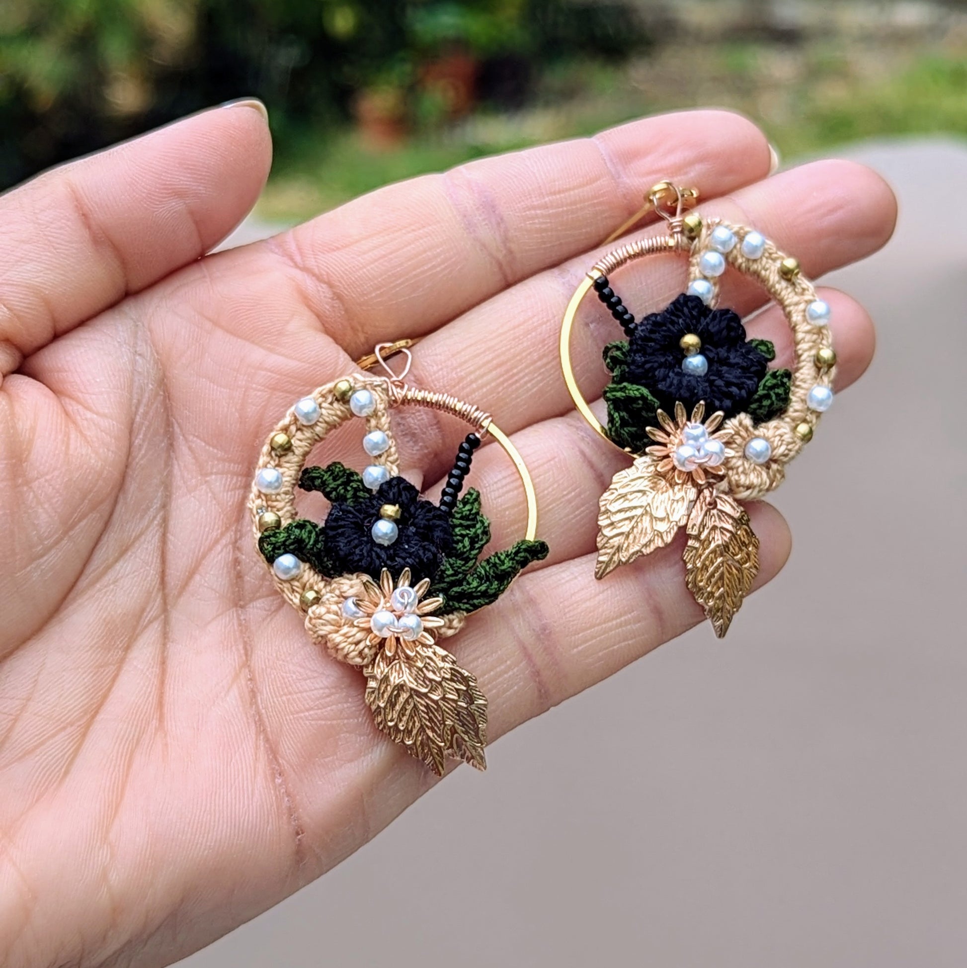Close-up Aloki floral hoop earrings in hand, highlighting black crochet, olive green leaves, and pearl accents.