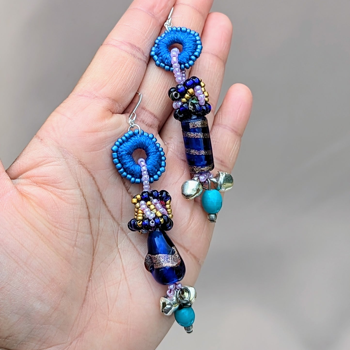 Hand holding blue asymmetric earrings showing mixed media beadwork and silver-plated hypoallergenic hooks.