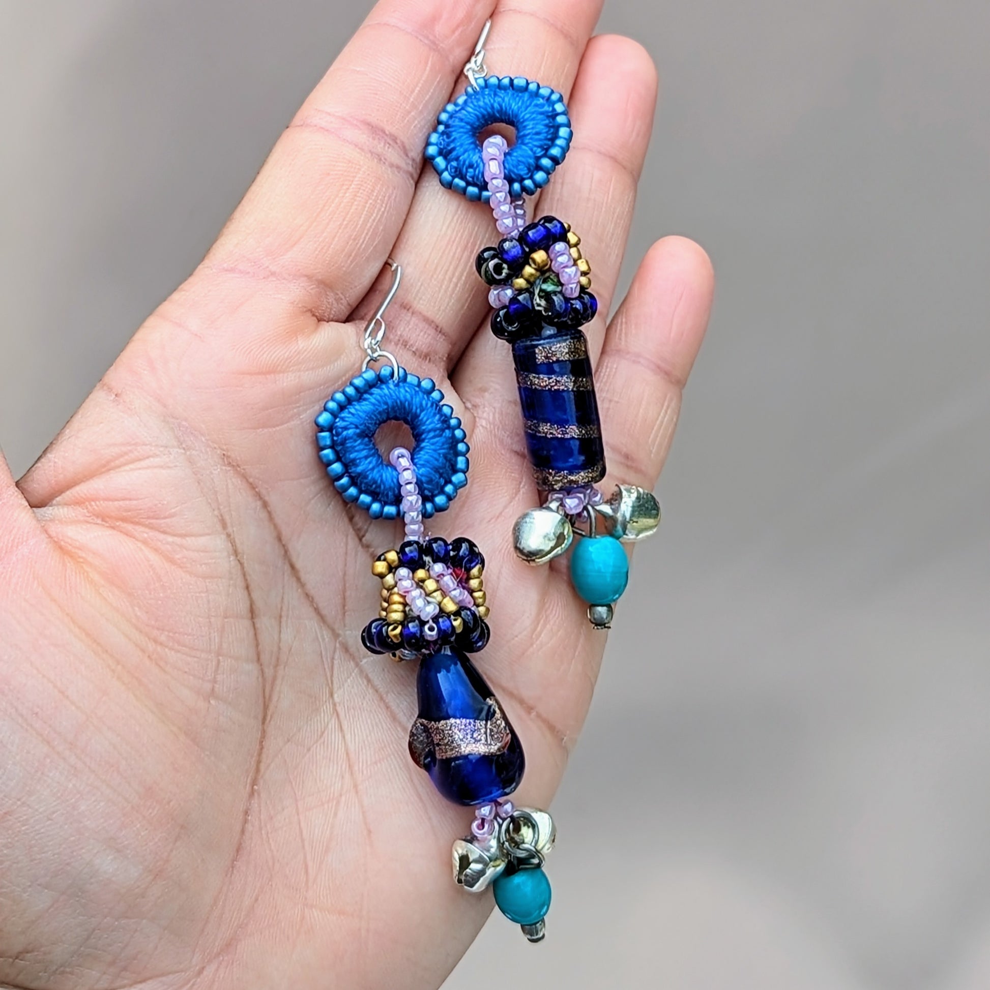 Hand holding blue asymmetric earrings showing mixed media beadwork and silver-plated hypoallergenic hooks.