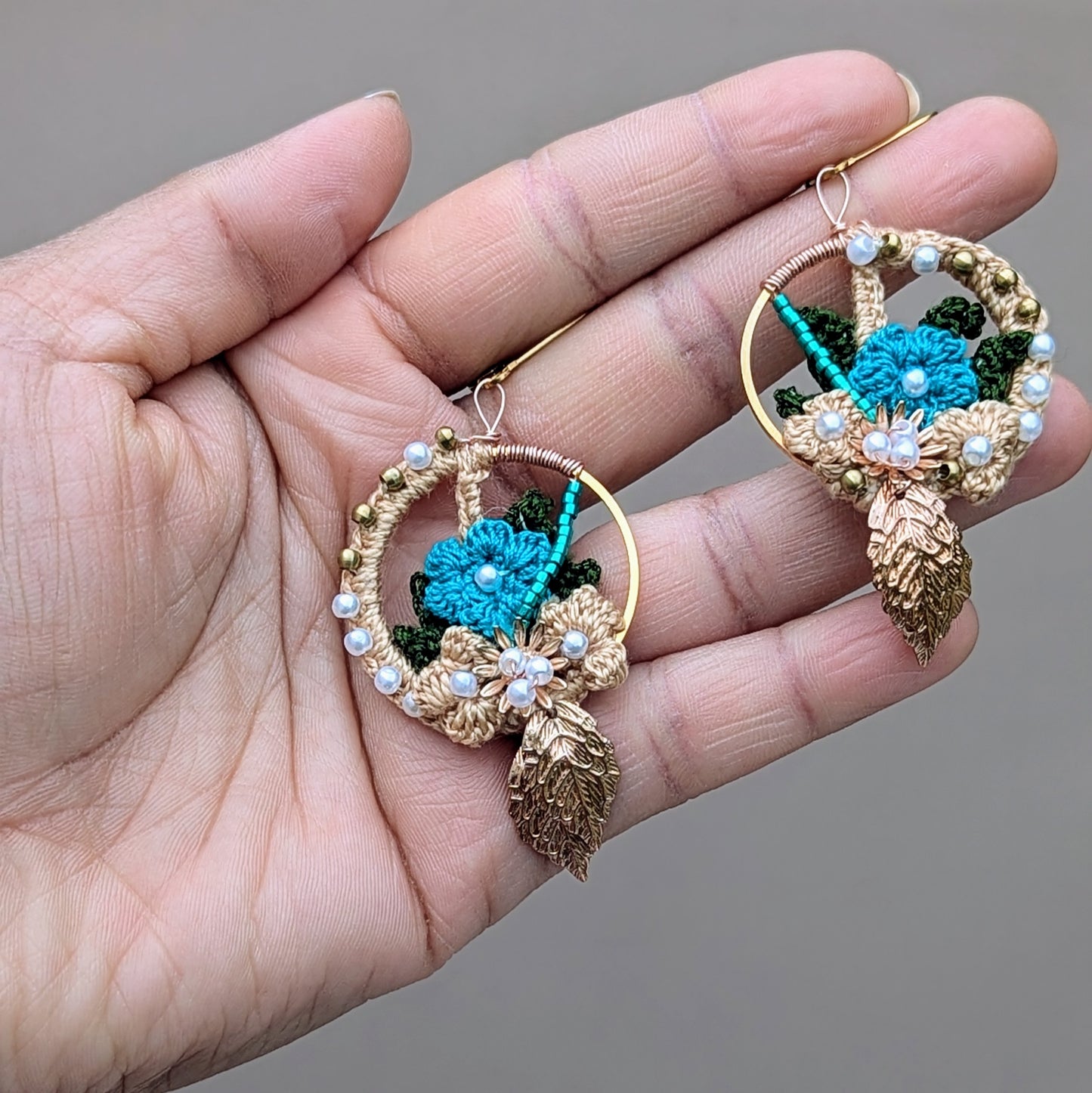 Hand holding a one-of-a-kind sustainably handcrafted pair of Aloki blue and gold hoop earrings inspired by Indian heritage, showcasing floral crochet with seed beads and golden leaf accents.