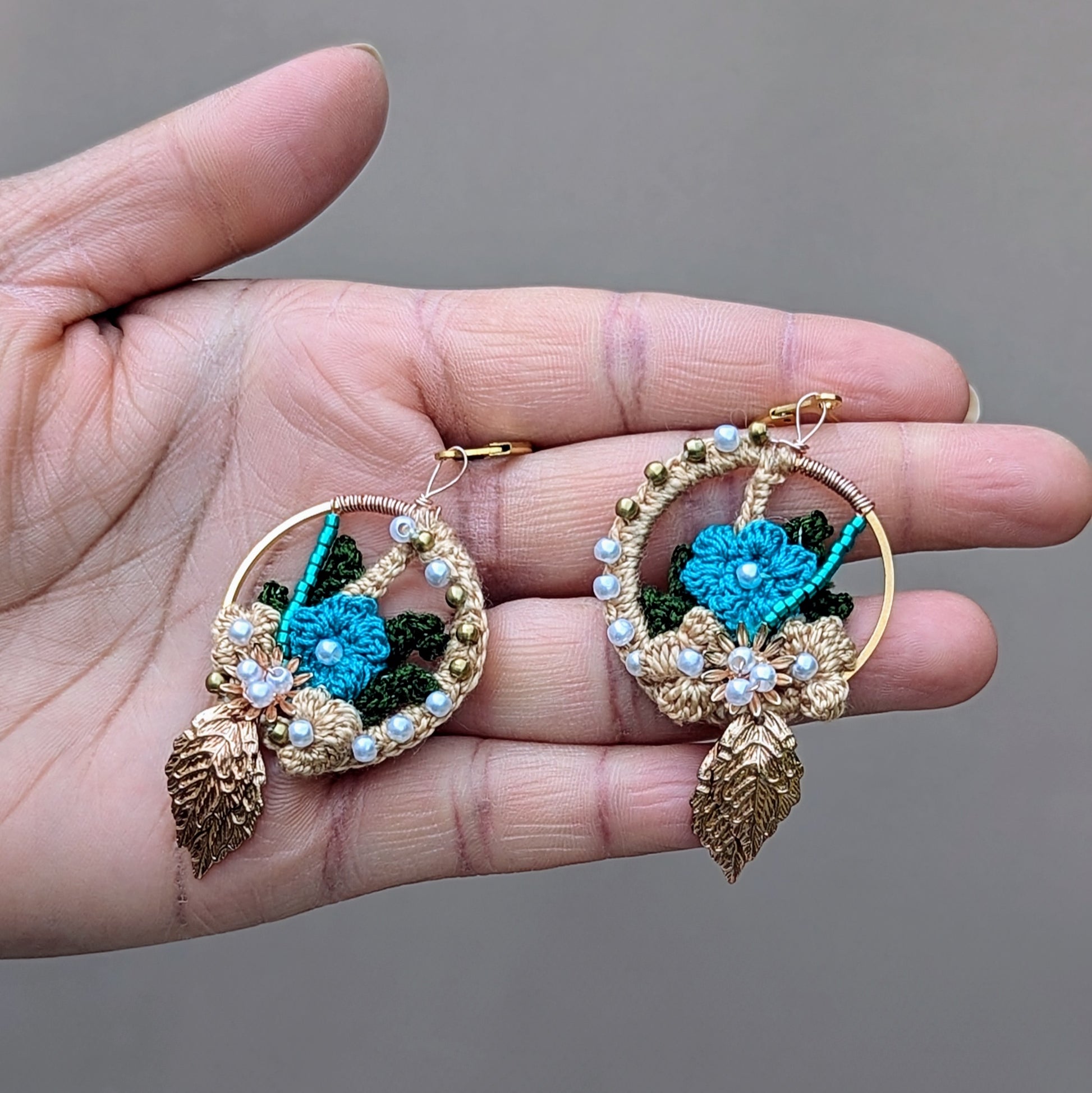 Hand holding a pair of statement Indian heritage inspired earrings with gold hoops, blue and green crochet accents, and seed pearl details. Exclusively created for and curated by Mayaani Jewellery UK.