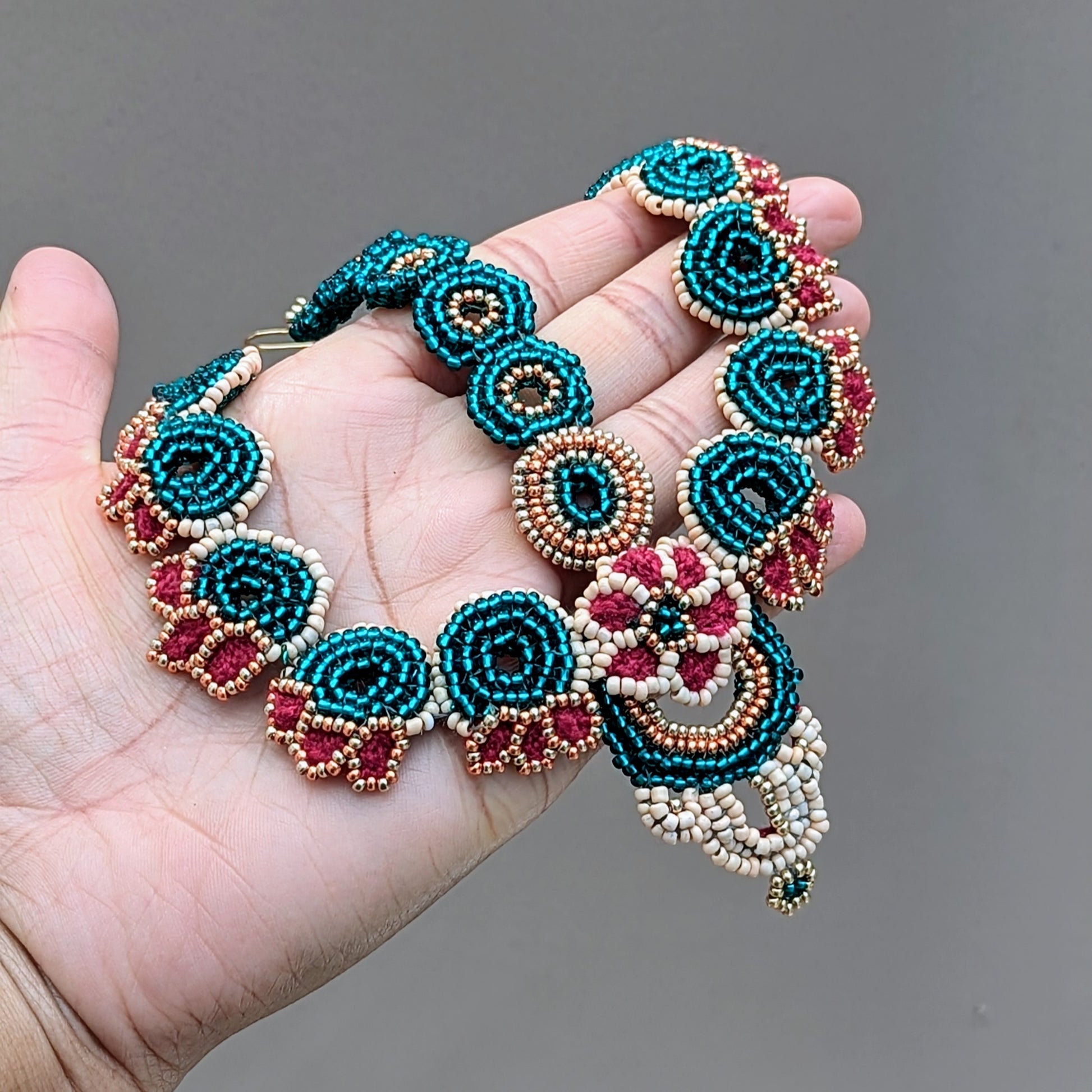 Close-up of hand holding the Neha Collection handmade Indian bridal headpiece (maang tikka) featuring emerald green, peach, red and gold beadwork in circular floral patterns, artisan crafted by Mayaani Jewellery UK.