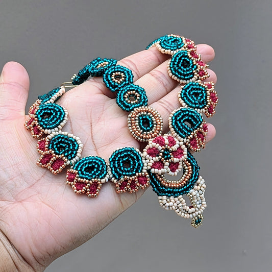 Close-up of hand holding the Neha Collection handmade Indian bridal headpiece (maang tikka) featuring emerald green, peach, red and gold beadwork in circular floral patterns, artisan crafted by Mayaani Jewellery UK.