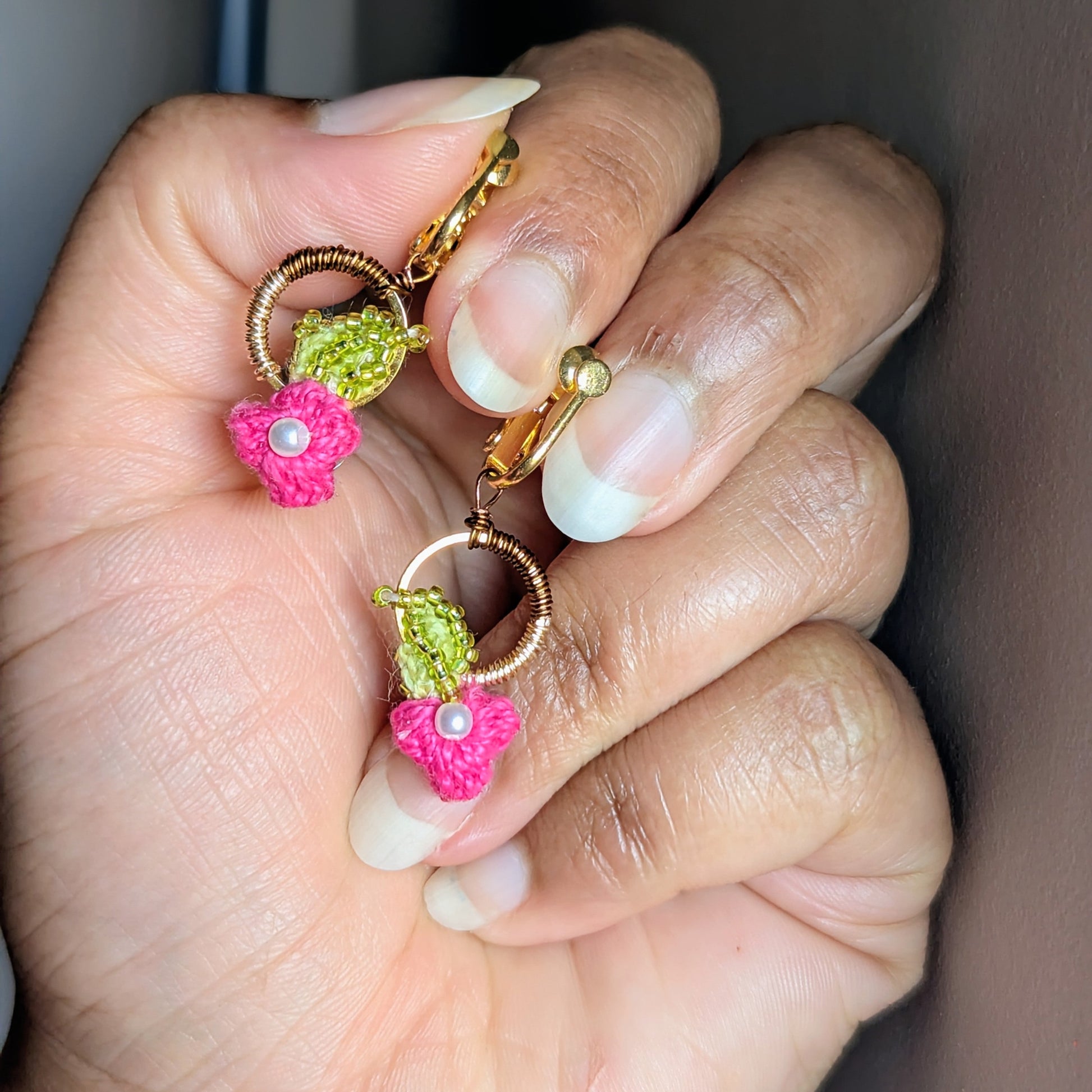 A close-up view of a hand holding the Pallavi pair of clip-on luxury handmade pink, chartreuse green, gold and pearl earrings designed for comfort for women with non-pierced ears.
