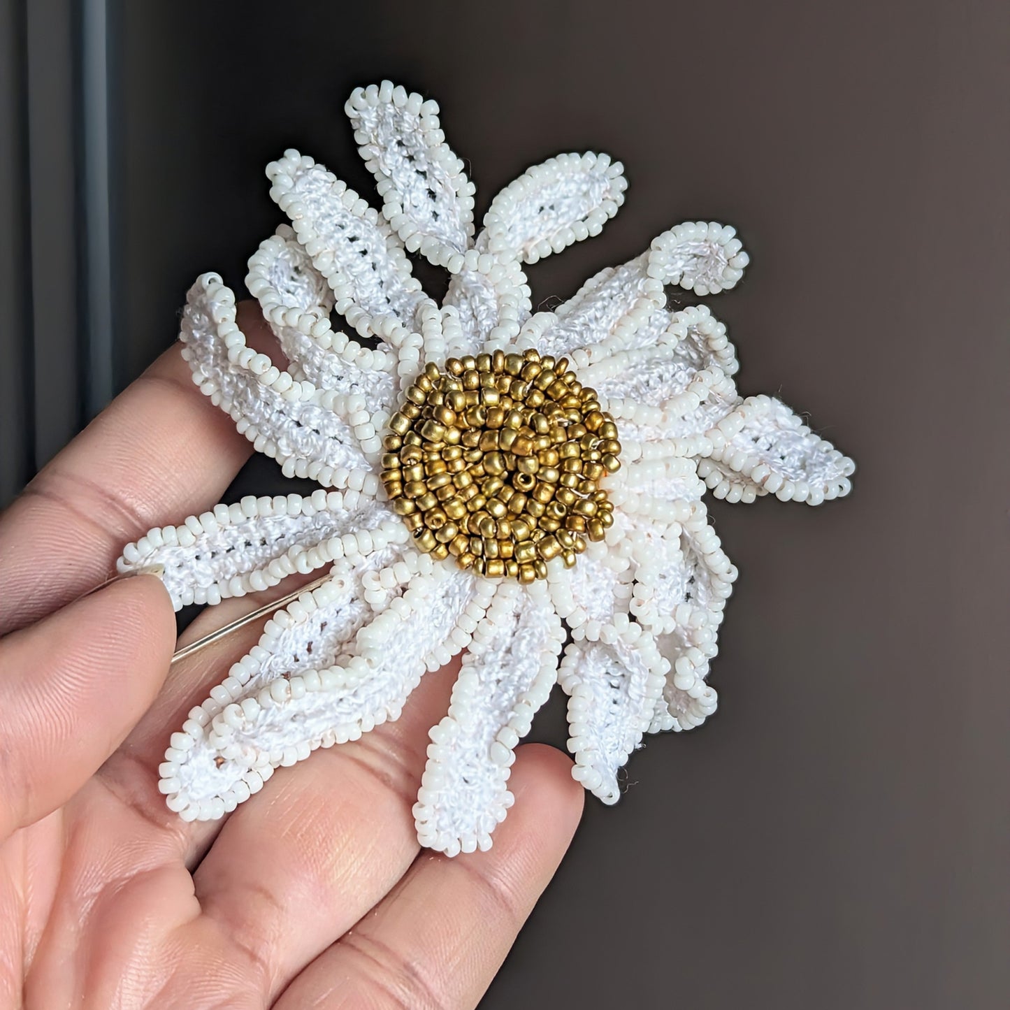 One of a kind Daisy floral inspired brooch held in hand against a dark grey backdrop showcasing crochet and beaded sculptural petals  hand made for Mayaani Jewellery UK.