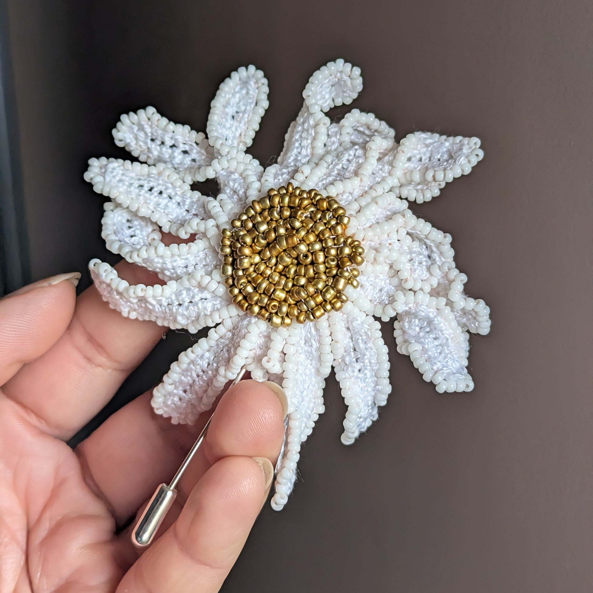 A botanical daisy flower inspired artisan made gold and white brooch pin which can be worn as a hat or lapel pin pictured in hand against a dark grey neutral backdrop.