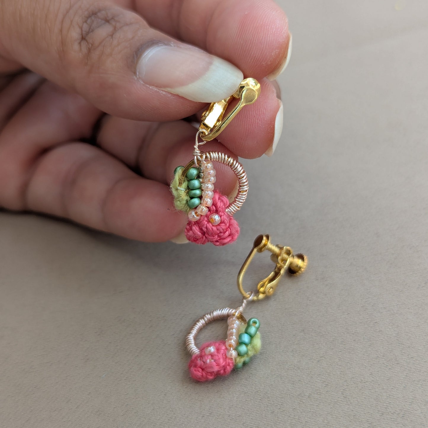 A close-up of a hand holding a pair of Indian hertiage inspired Anandi hand woven gold hoop earrings with pink and green beaded botanical details against a neutral beige background.