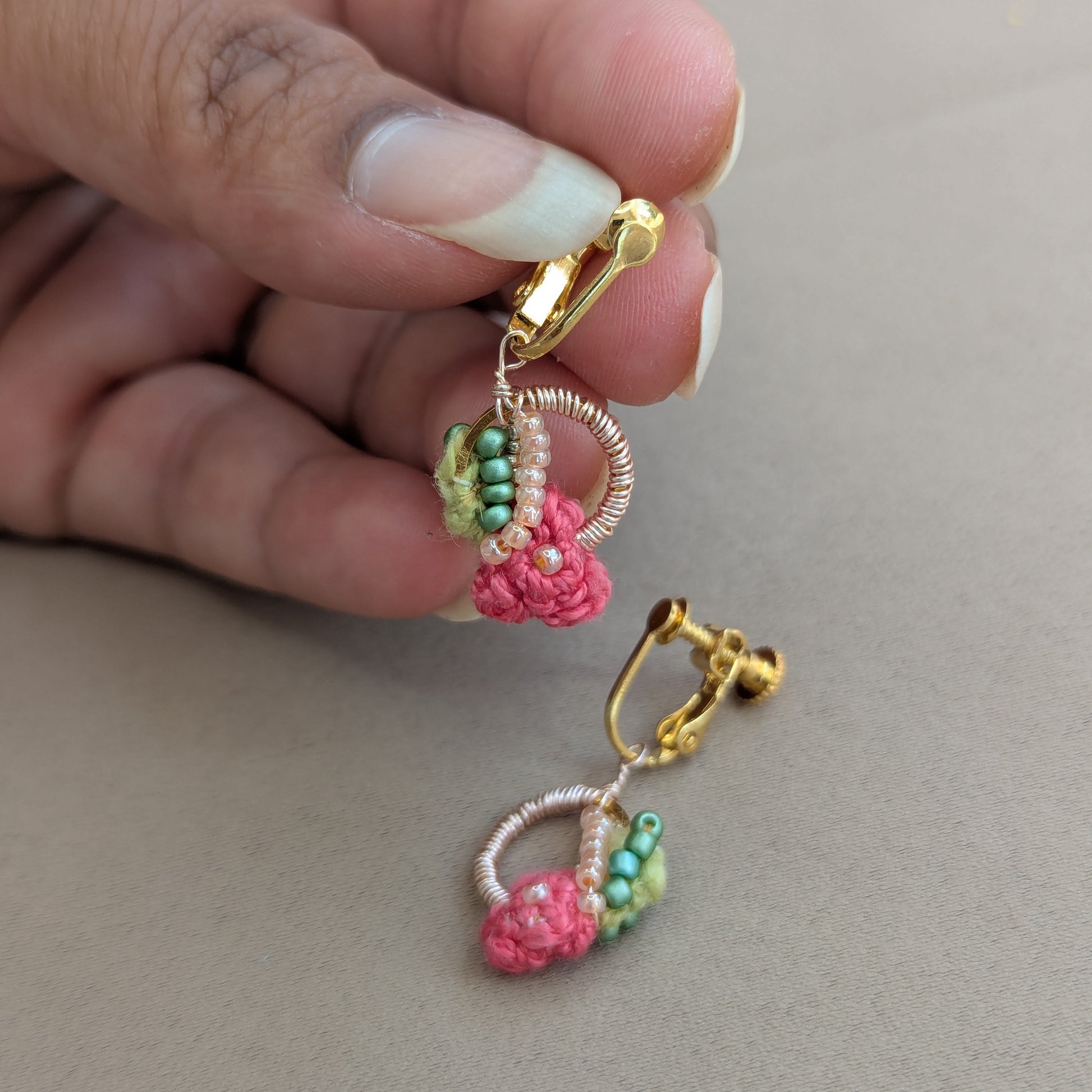 A close-up of a hand holding a pair of Indian hertiage inspired Anandi hand woven gold hoop earrings with pink and green beaded botanical details against a neutral beige background.