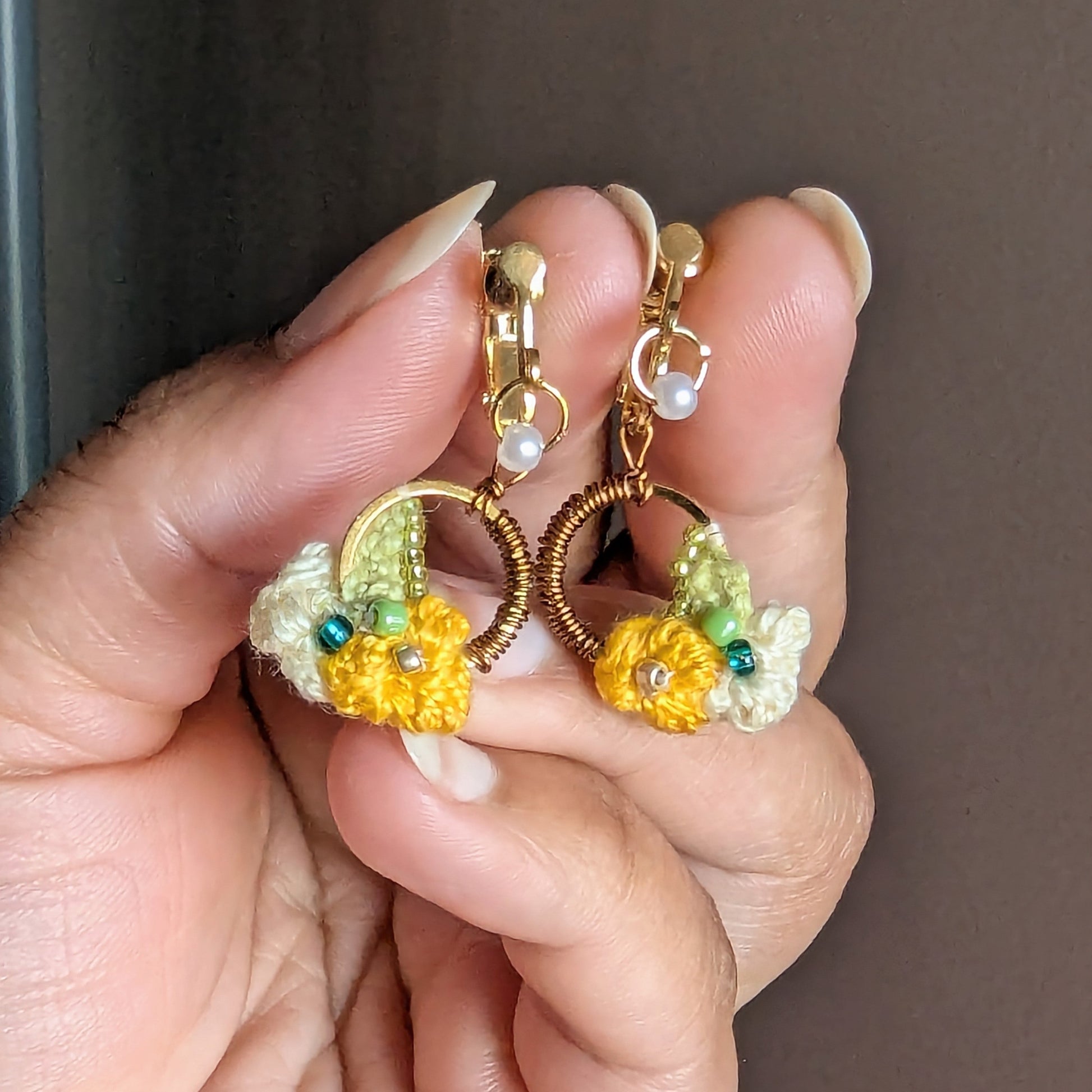 Hand holding a unique lightweight pair of Sauri yellow, cream, green, gold, bronze and pearl botanical inspired clip-on earrings sustainably handmade at Mayaani Jewellery UK using a combination of hand wire wrapping, hand bead embroidery and hand crochet.