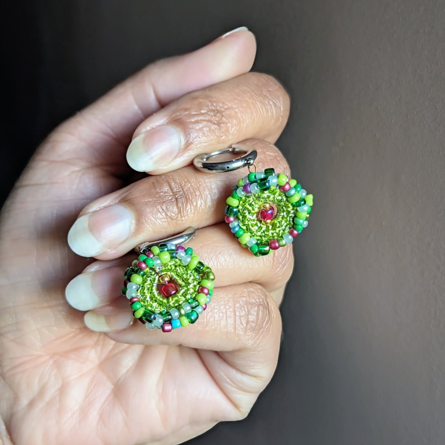 Vana garden-inspired handmade earrings held in hand, highlighting intricate bead crochet circles in green, red, blue, and purple tones with delicate stainless steel hoops.