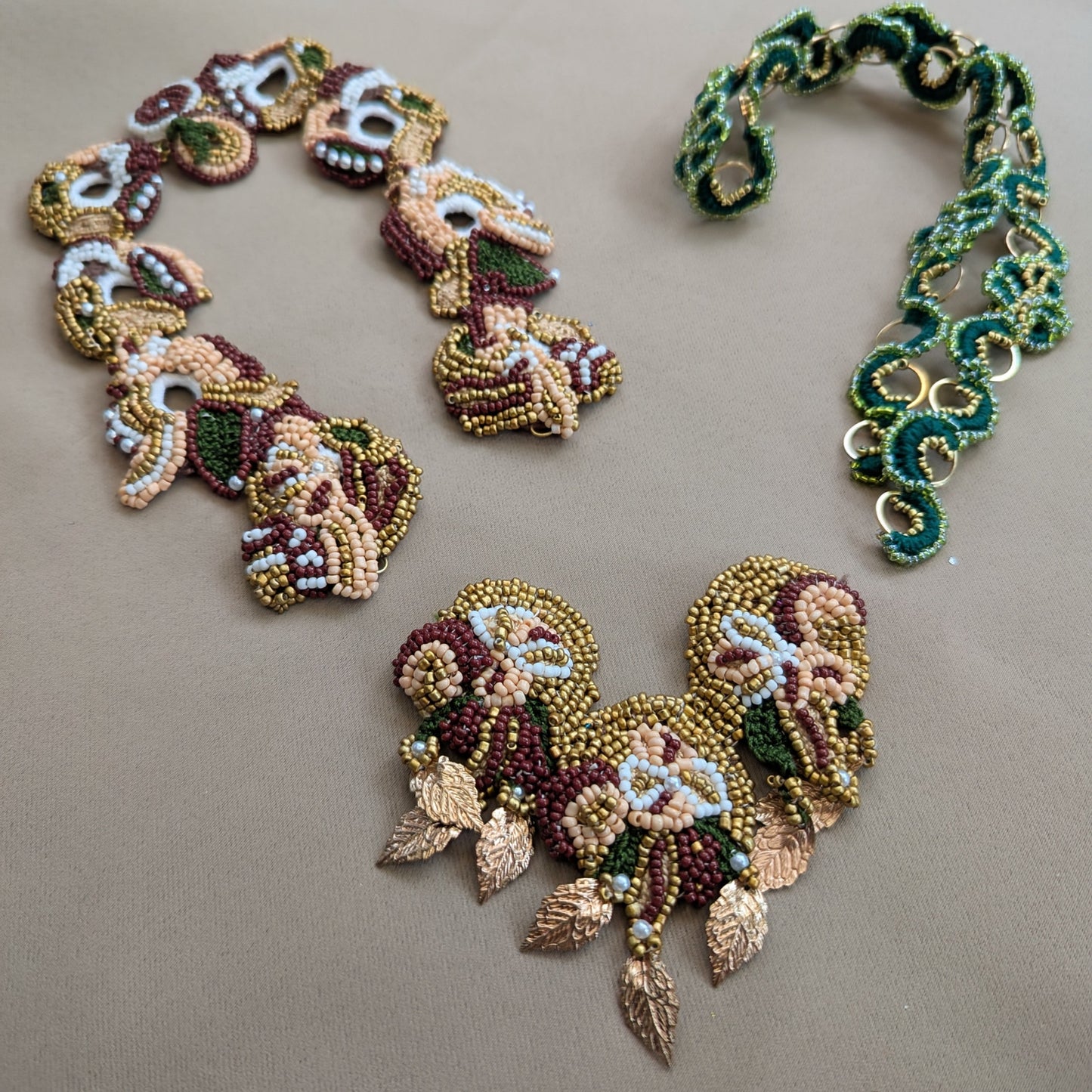 Three handmade beaded jewellery pieces, including a necklace, bracelet, and earrings, featuring intricate floral and skull motifs in gold, green, and burgundy beads, displayed on a neutral beige surface.