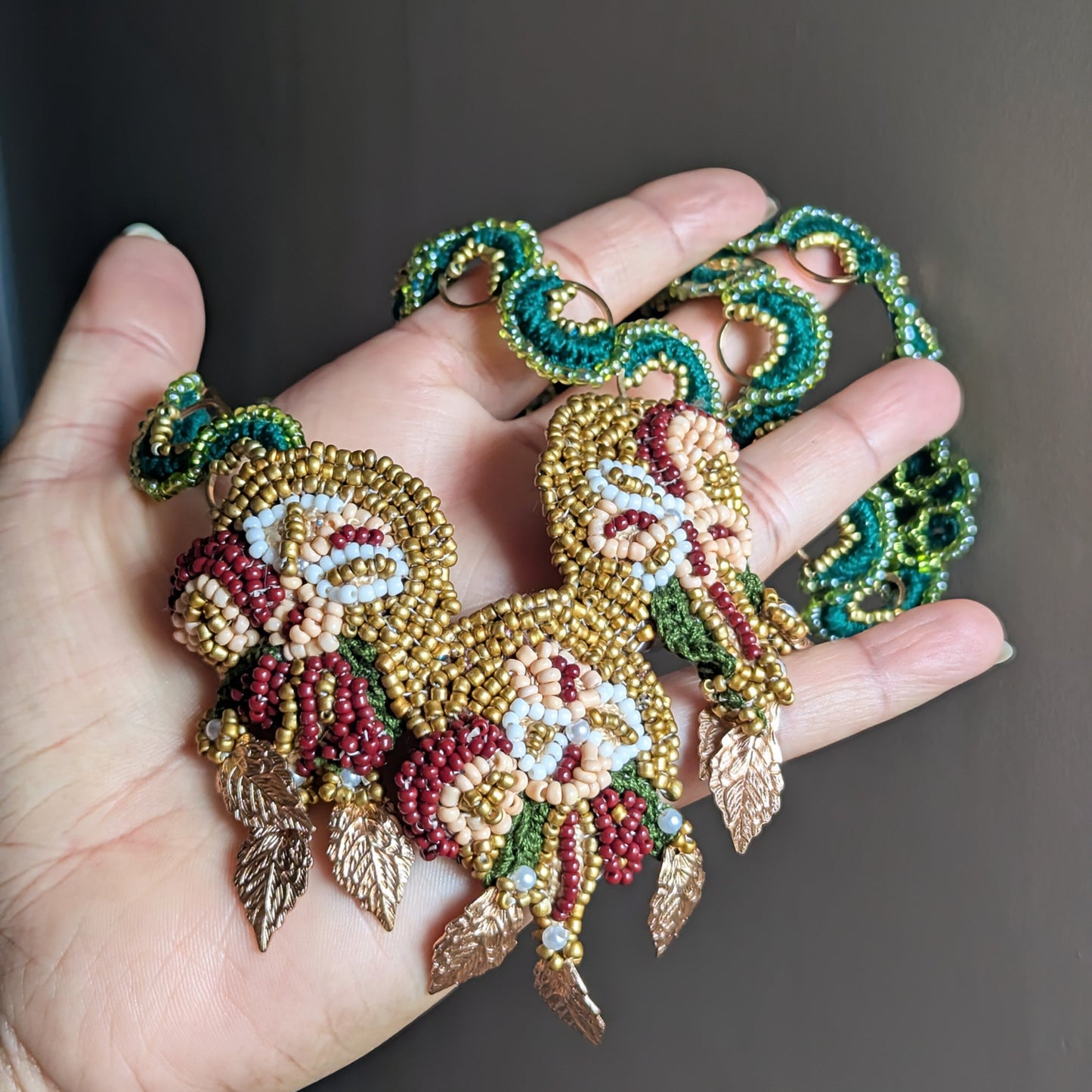 Hand held view of the artisan bead woven Aishwarya green, gold and red statement pendant inspired by florals attached to a custom made emerald green and gold chain.