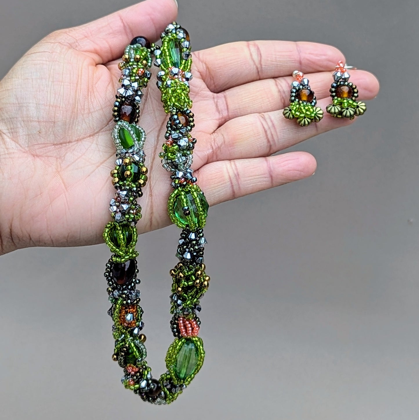Hand holding an artisanal green gold and brown beaded necklace and earrings against a neutral background.