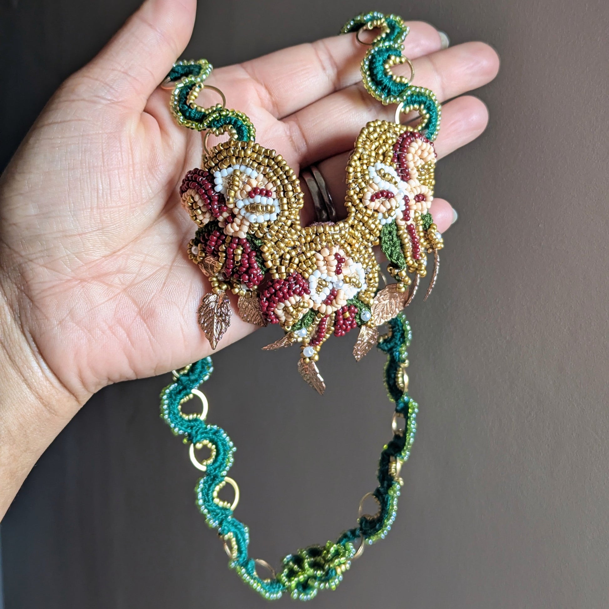 A hand holding the Aishwarya couture handmade beaded Indian inspired necklace featuring a central floral design with gold, red, and white beads, and a green and gold beaded chain.