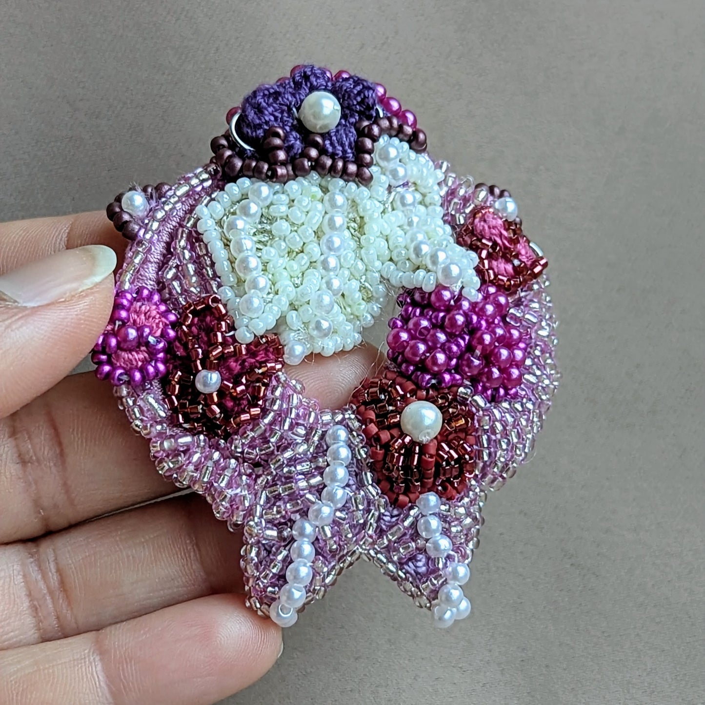 Hand holding the intricately beaded detachable pendant from the 'Gulab Rani' Indian bridal necklace handcrafted by Mayaani Jewellery UK for the modern South Asian bride with an appreciation for Western aesthetics. Featuring rich, regal tones of purple, pink, red and ivory displayed against a neutral background.