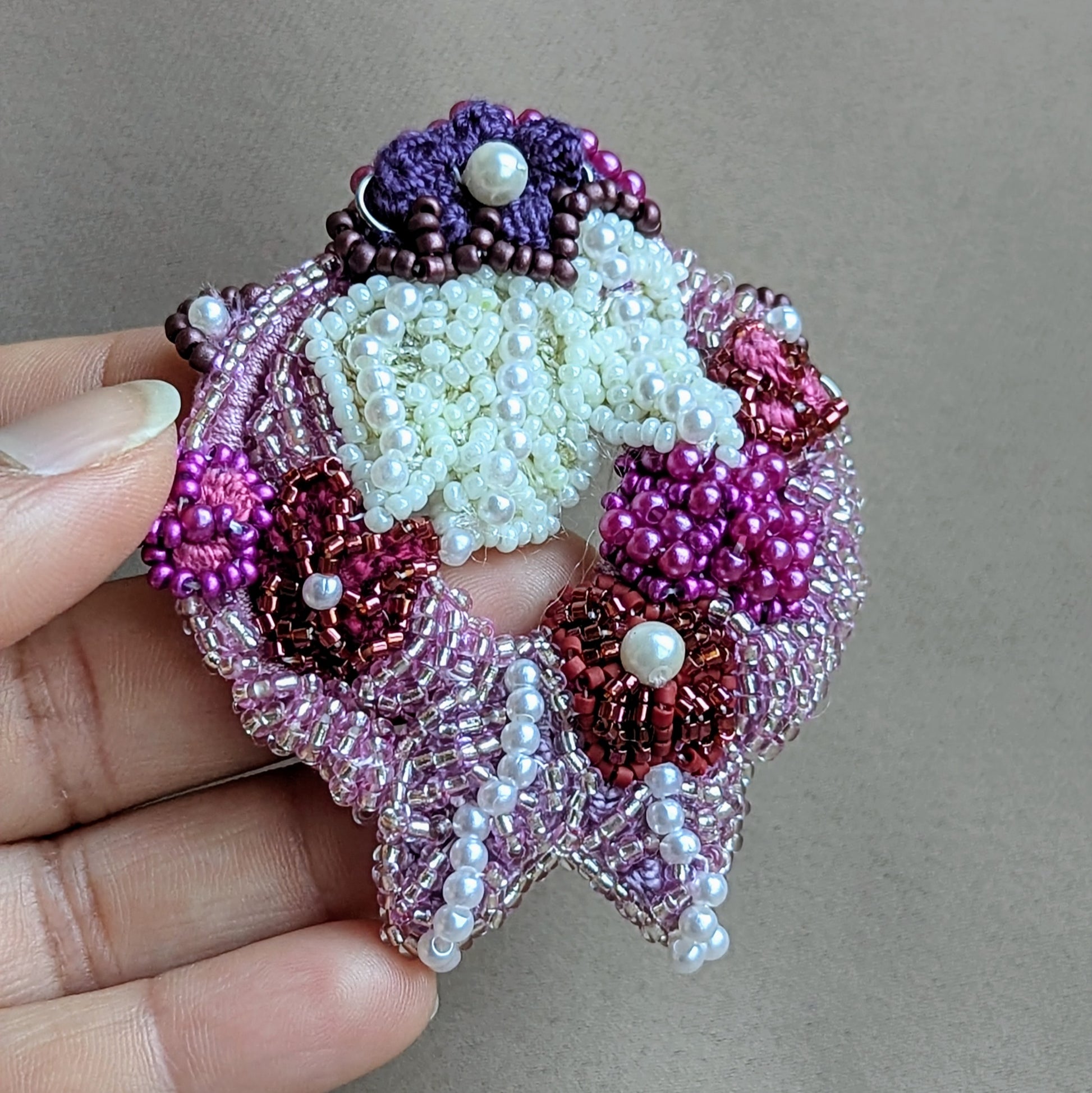 Hand holding the intricately beaded detachable pendant from the 'Gulab Rani' Indian bridal necklace handcrafted by Mayaani Jewellery UK for the modern South Asian bride with an appreciation for Western aesthetics. Featuring rich, regal tones of purple, pink, red and ivory displayed against a neutral background.