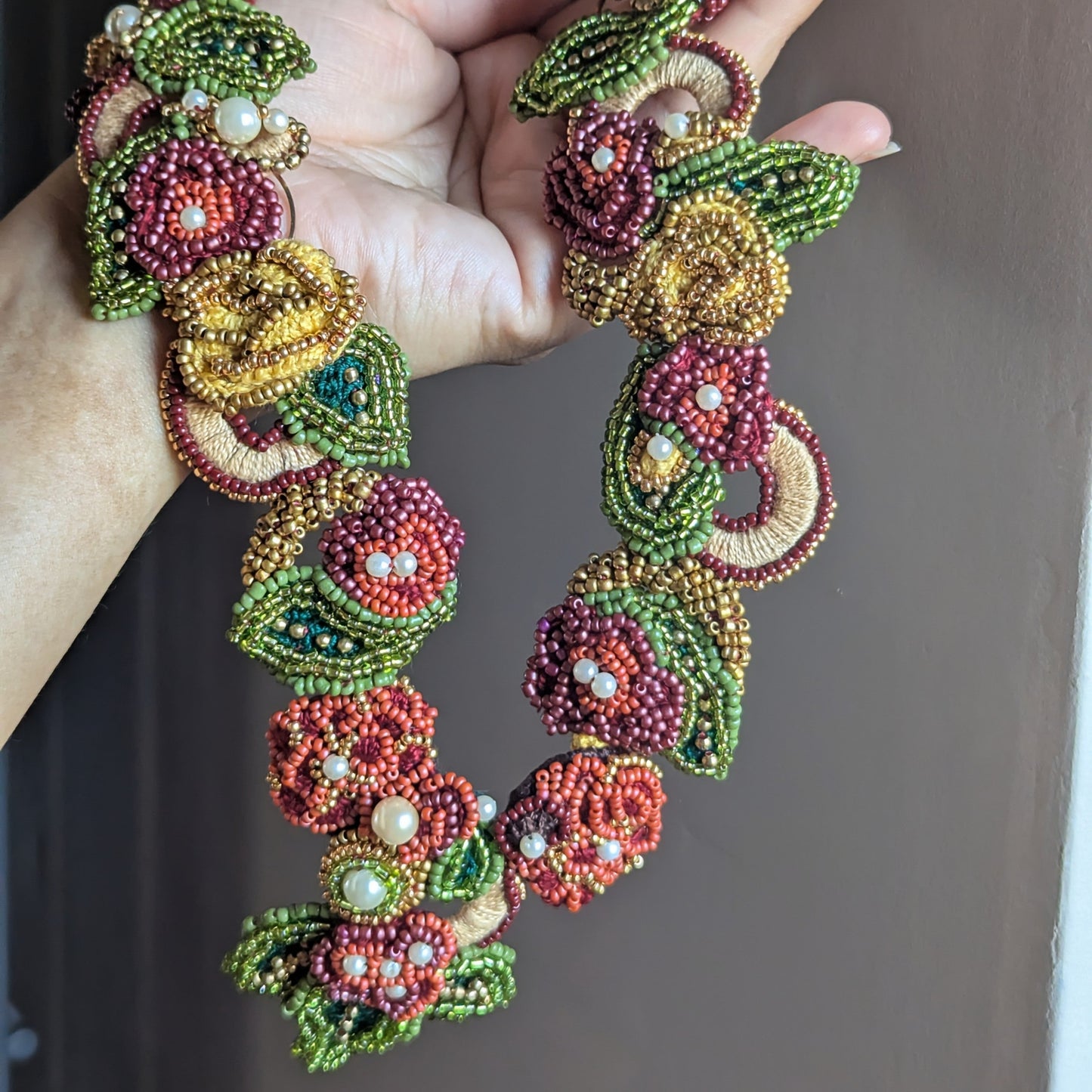 Hand holding a richly detailed burgundy, olive green, emerald green, gold and yellow modern fusion necklace sustainably handmade at Mayaani Jewellery UK and designed to offer two unique looks in one piece.