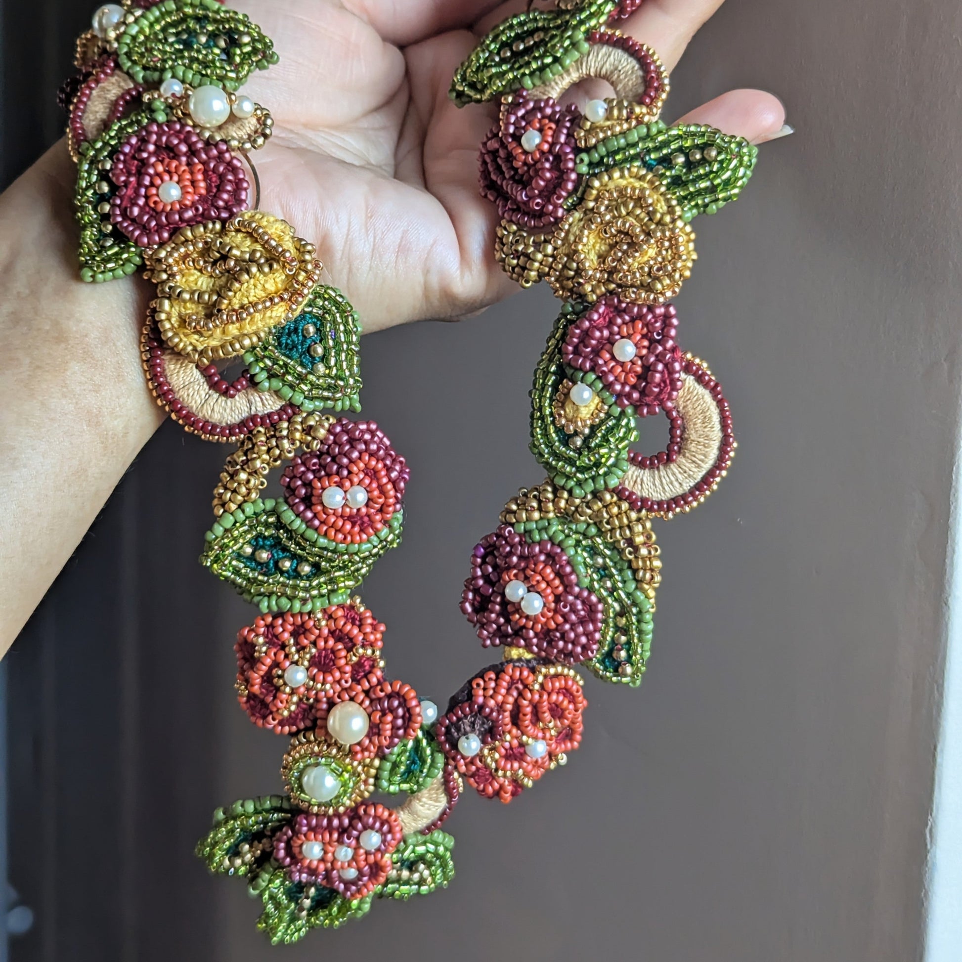 Hand holding a richly detailed burgundy, olive green, emerald green, gold and yellow modern fusion necklace sustainably handmade at Mayaani Jewellery UK and designed to offer two unique looks in one piece.