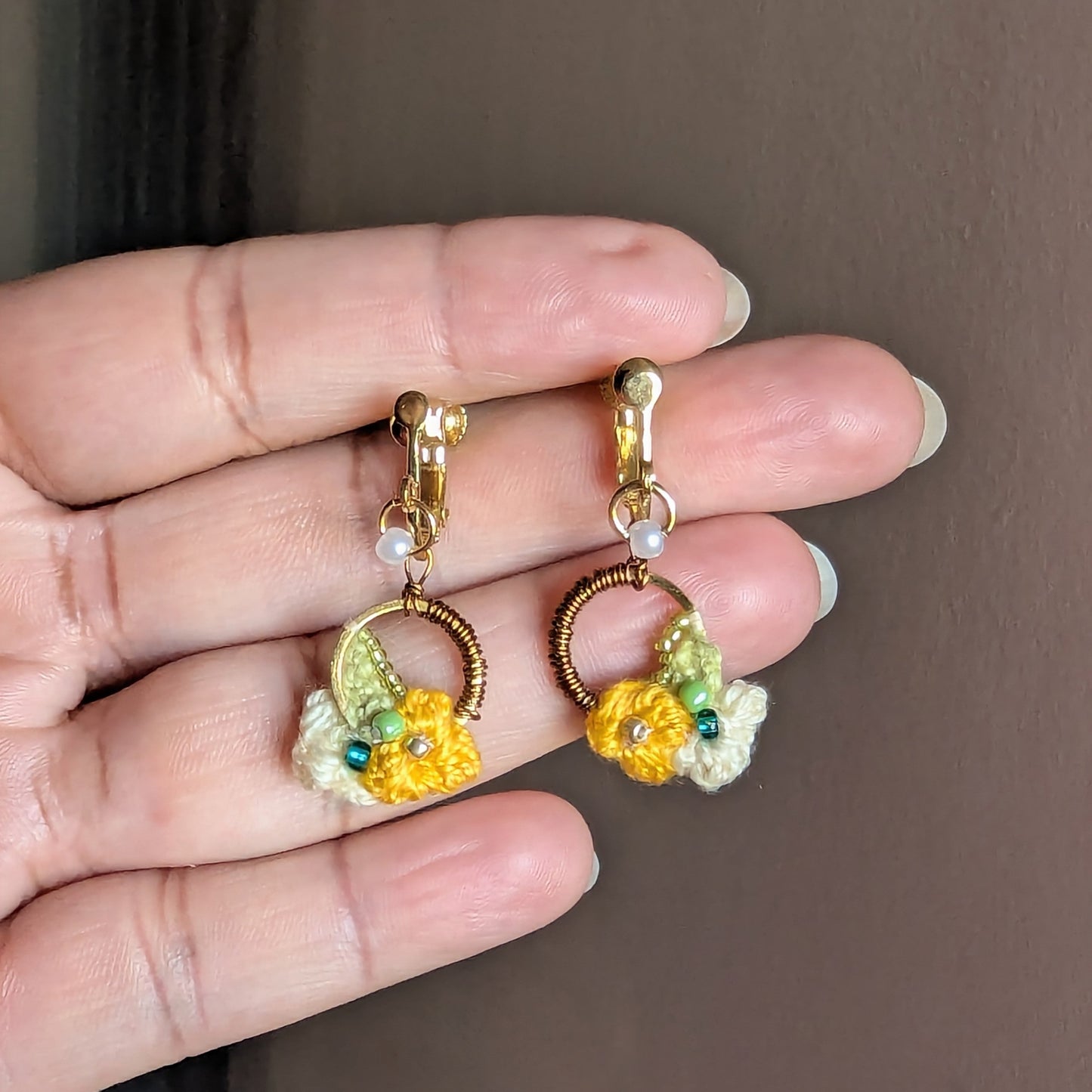 Hand held view of a one off pair of designer clip-on Sauri floral botanical inspired Indian fusion earrings in yellow, green, gold, bronze and pearl.